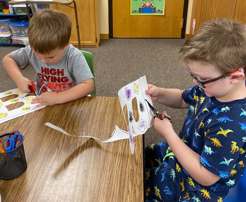 Two children cutting paper at a table in a classroom