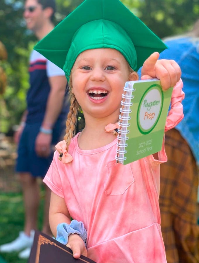 Child in a green graduation cap holding a small notebook with "Ragan Prep" on it