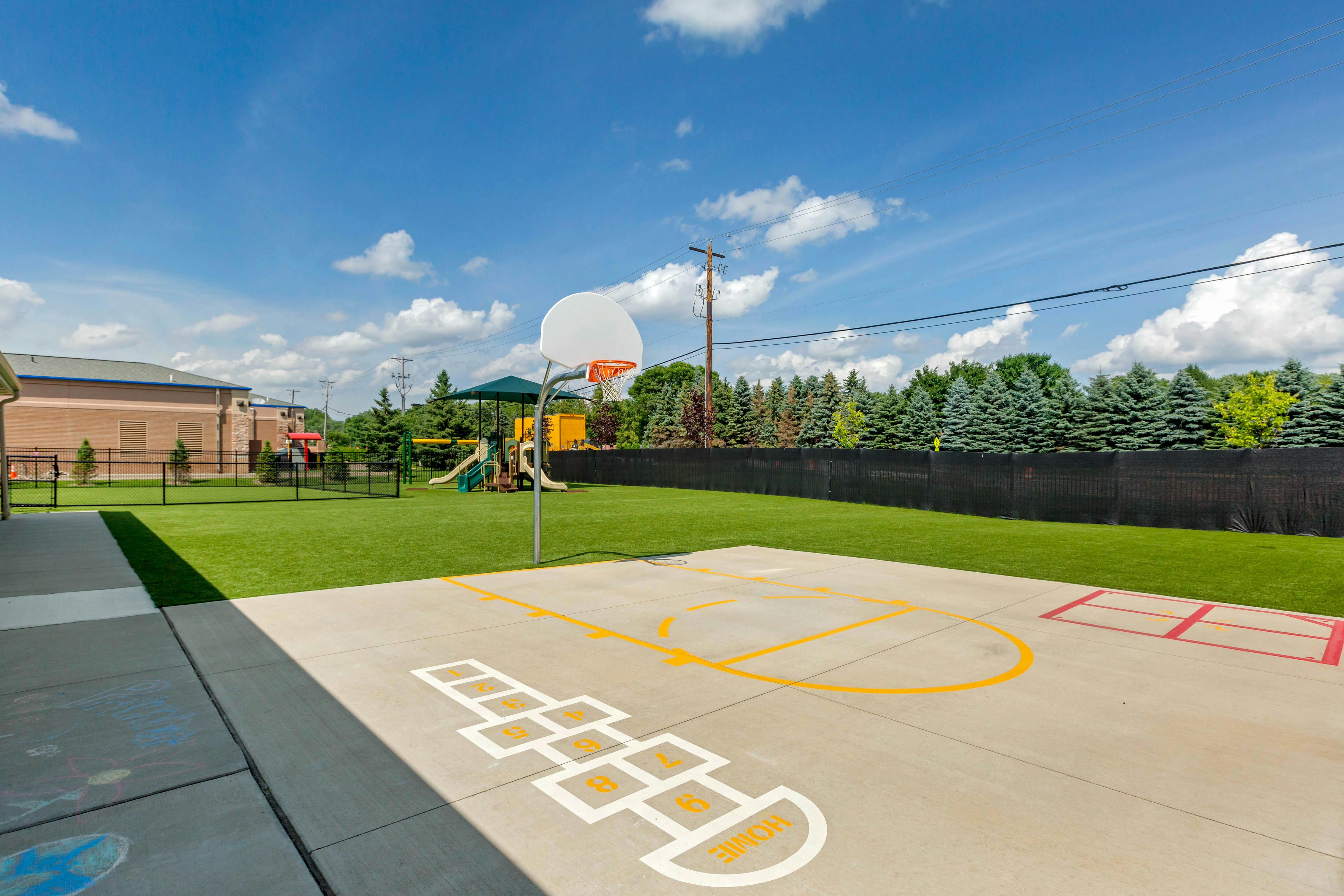 Outdoor playground area with a basketball hoop, hopscotch, and green turf