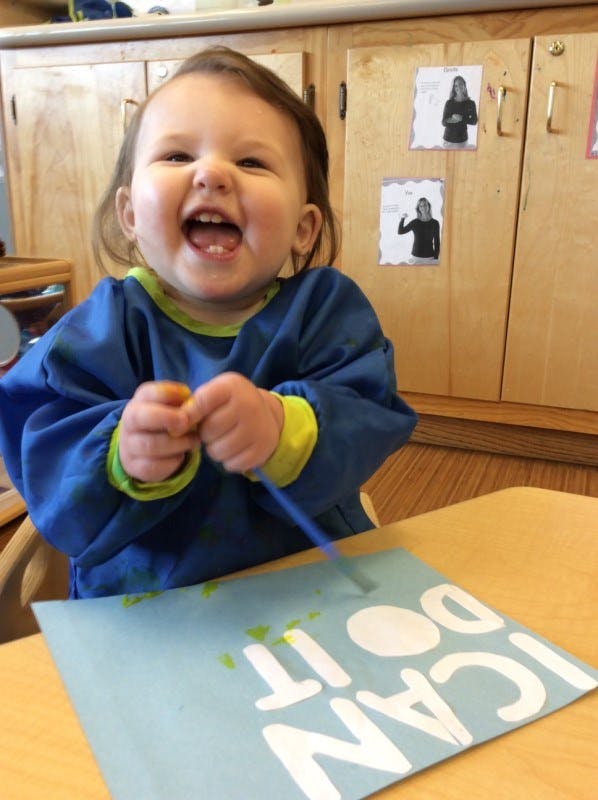 Child in a blue smock painting a paper with "I CAN DO IT" written on it