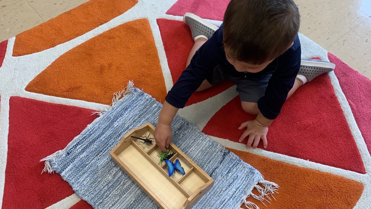Child on a colorful rug reaching for items in a wooden tray
