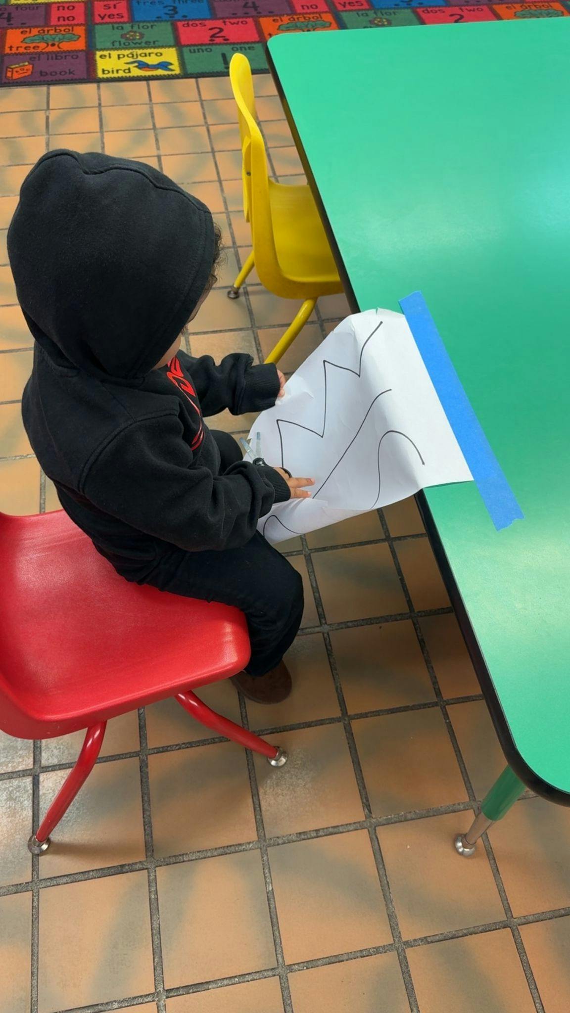 Child in a black hoodie holding a paper with wavy lines at a green table