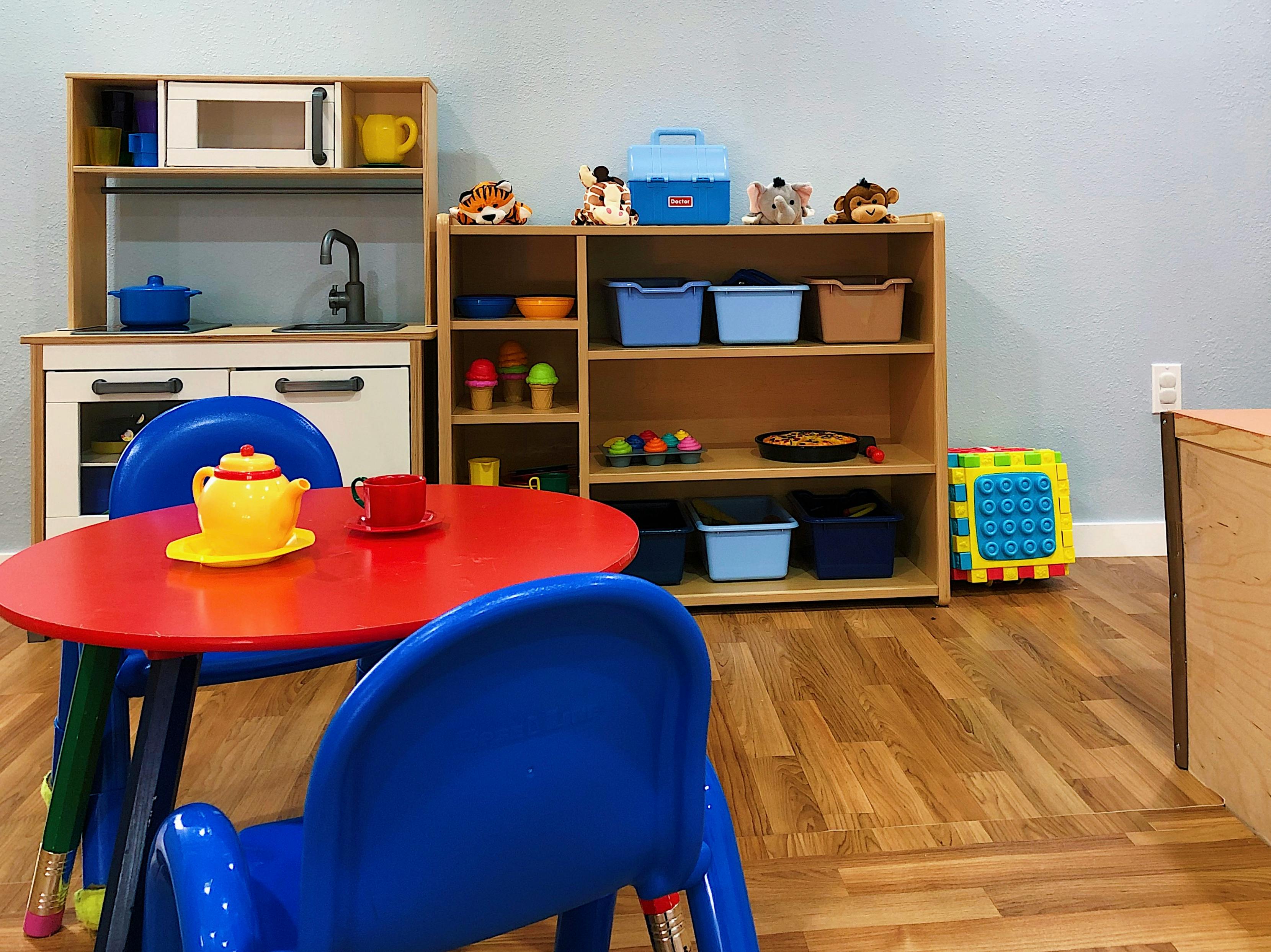 Play kitchen and shelves with toys in a room with a red table and blue chairs