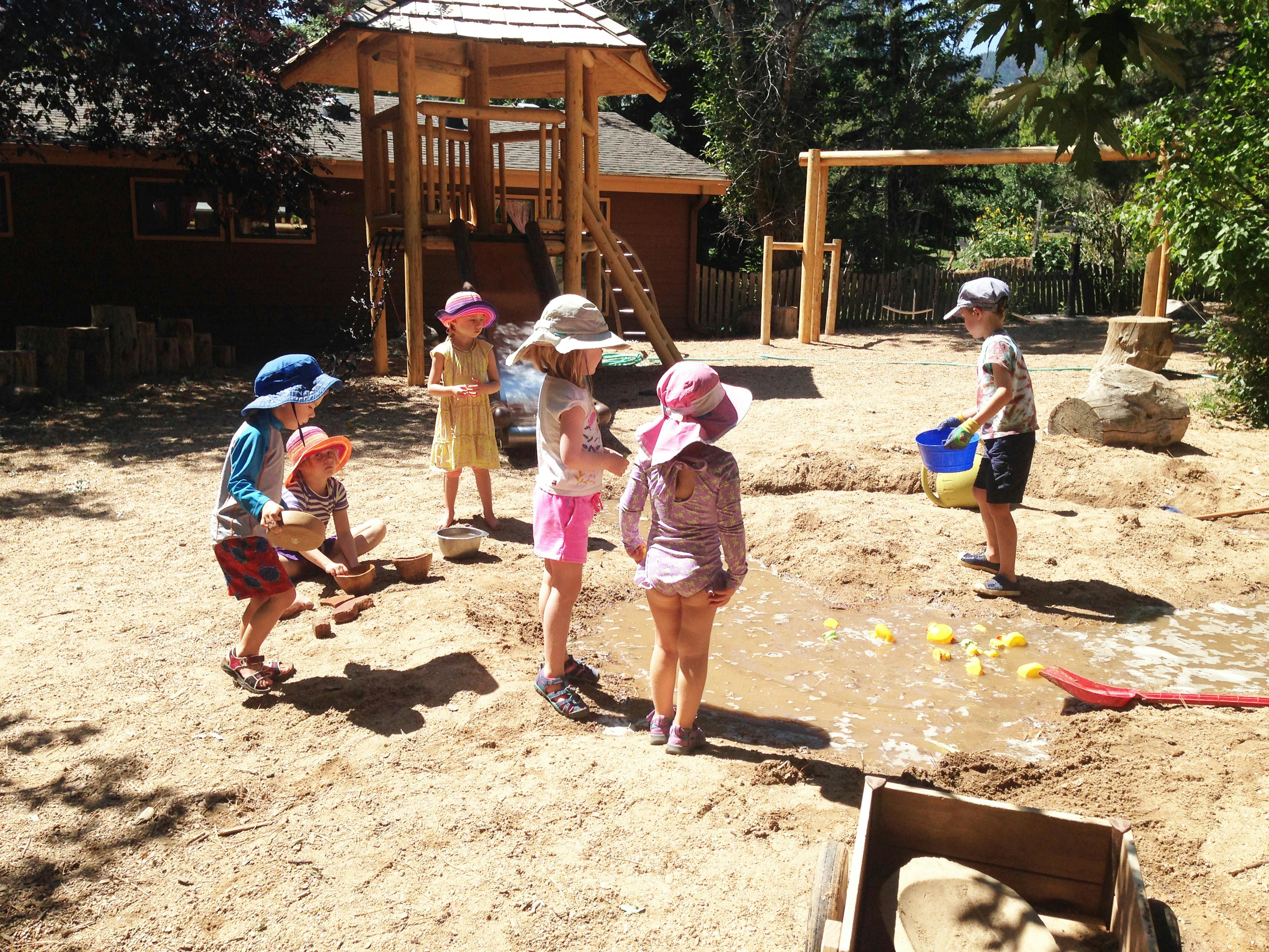 Children playing in a muddy area with a wooden play structure in the background