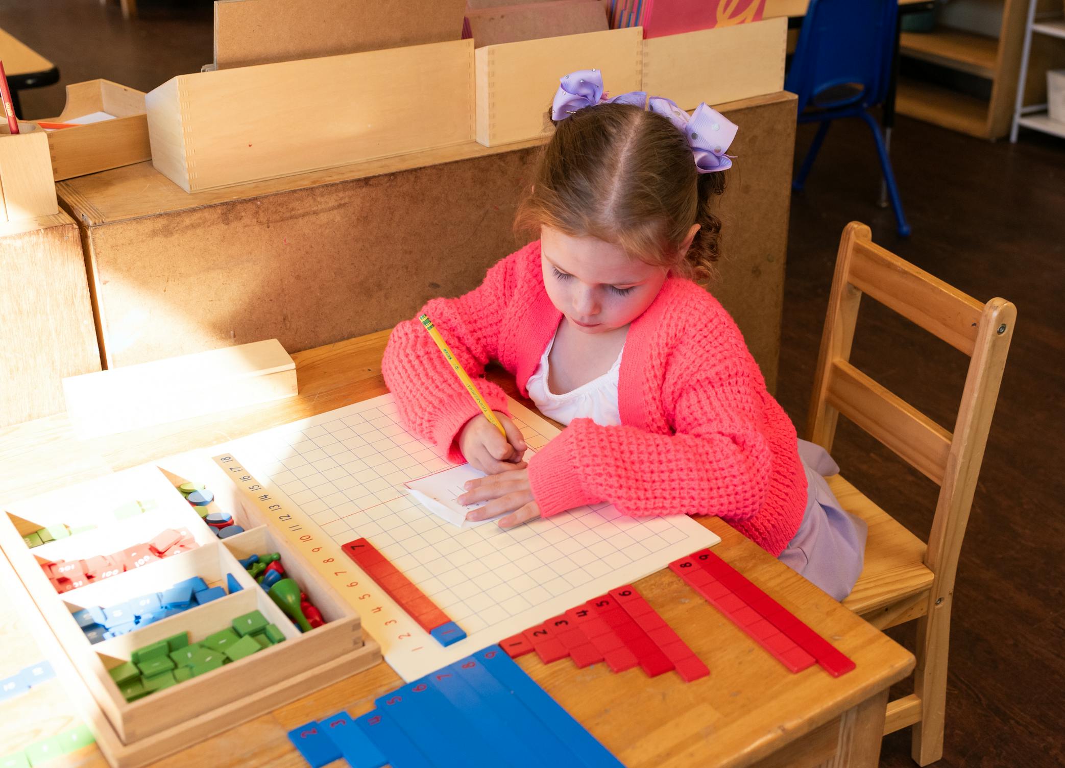 Child writing with a pencil on a grid worksheet at a table with math manipulatives