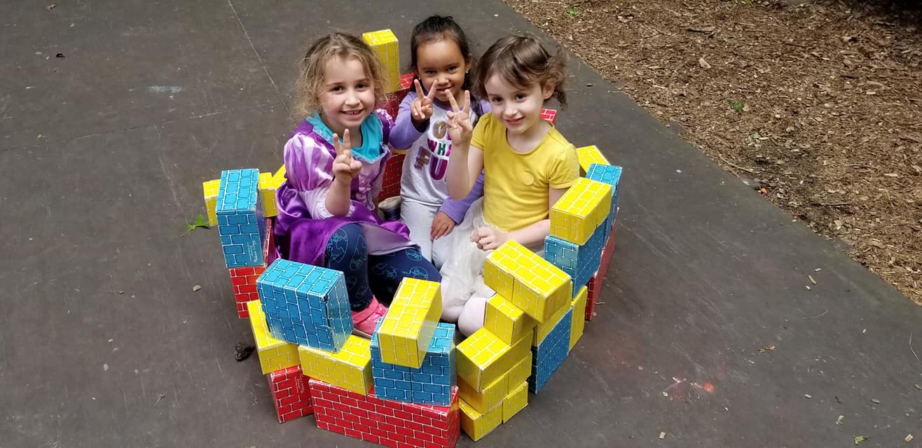 Three children sitting inside a structure made of large building blocks