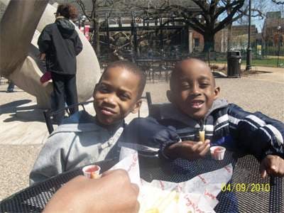 Two children sitting at a table outdoors, one holding a french fry