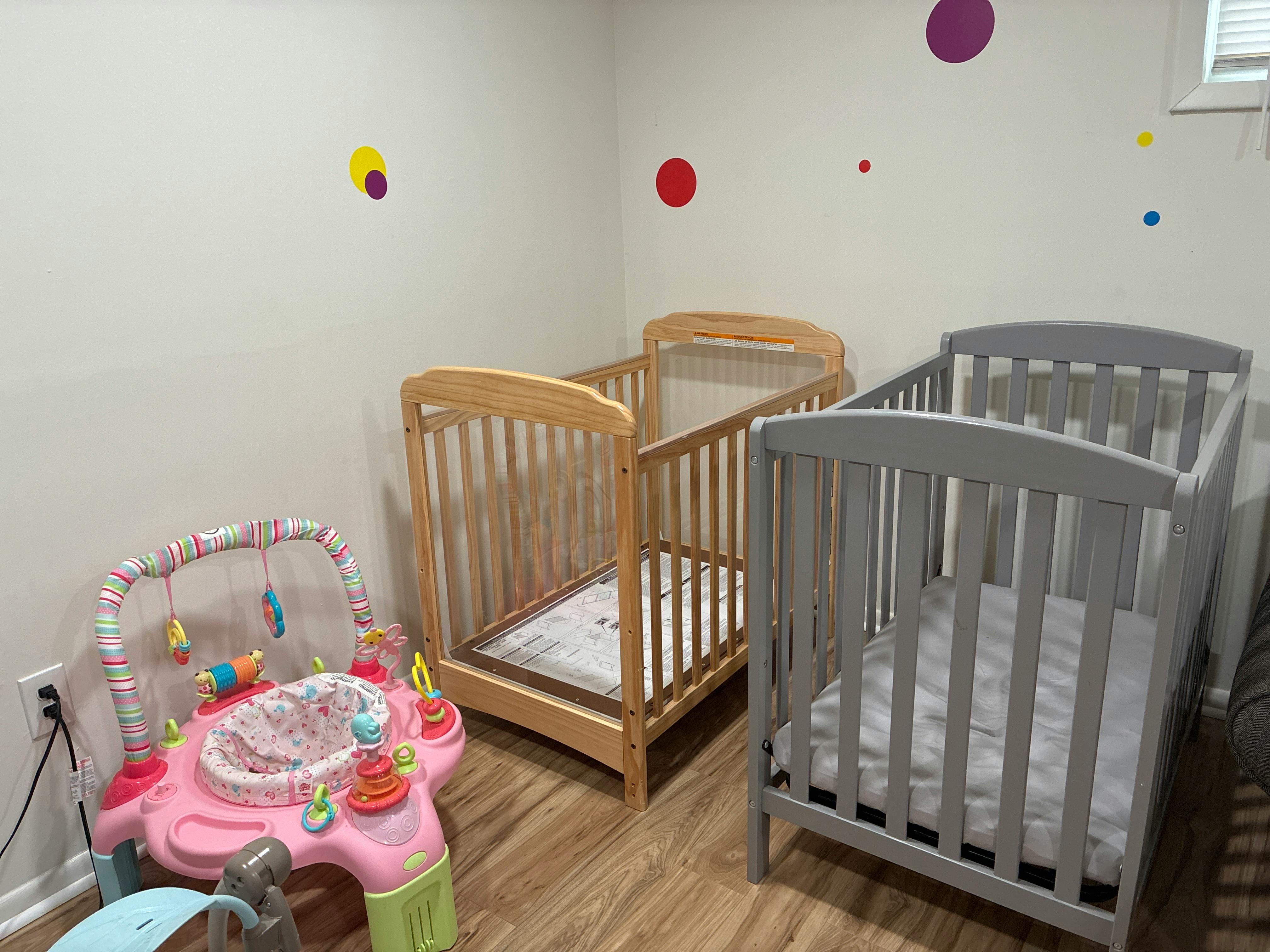 A pink infant activity center next to two baby cribs, one light wood and one grey