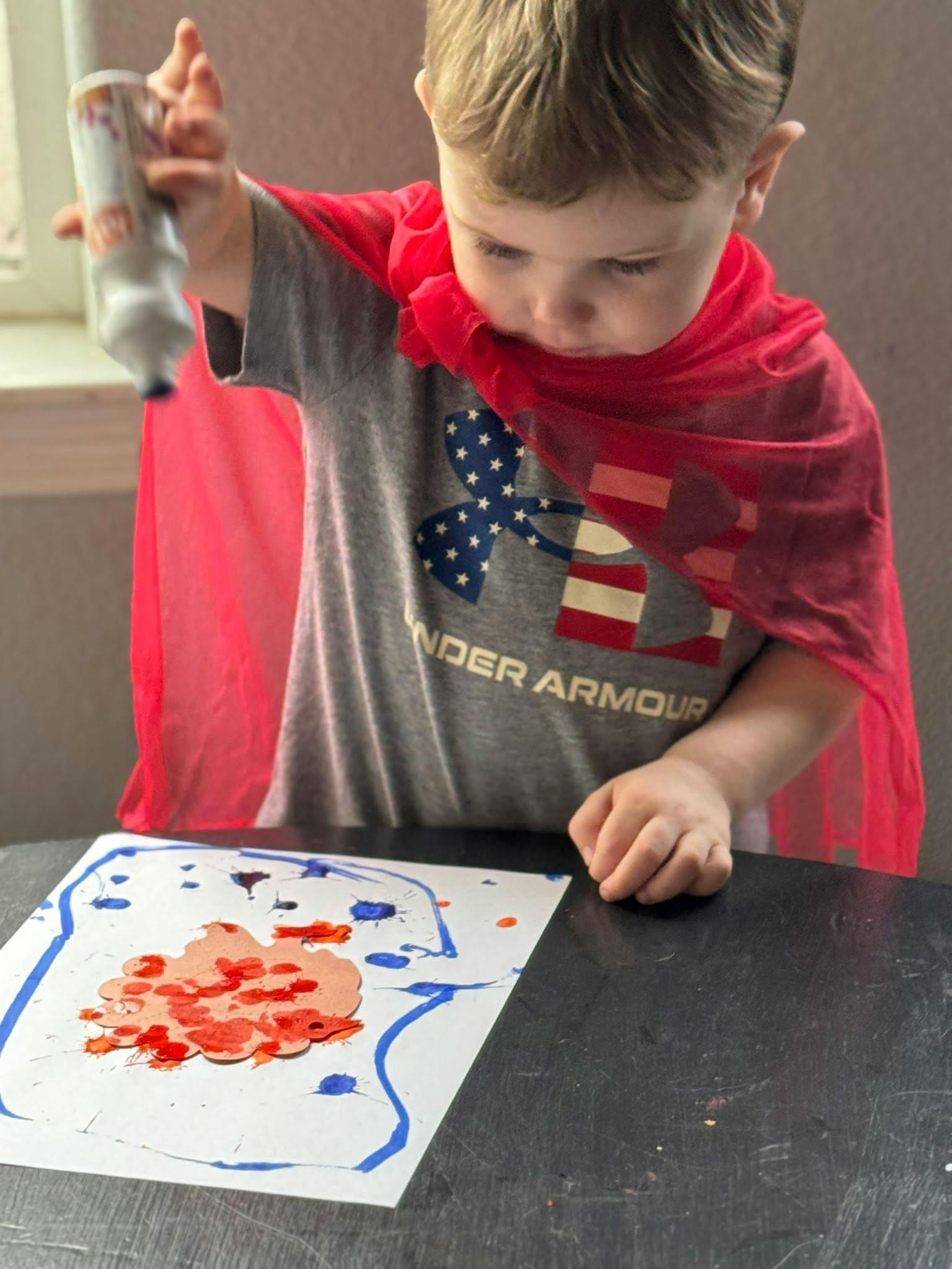 Child in a red cape squeezing paint onto paper