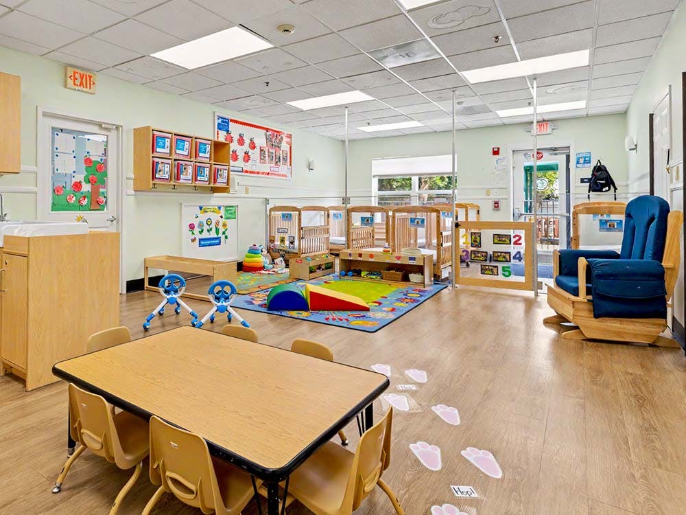 A classroom with small tables and chairs, cribs, a rocking chair, and playmats