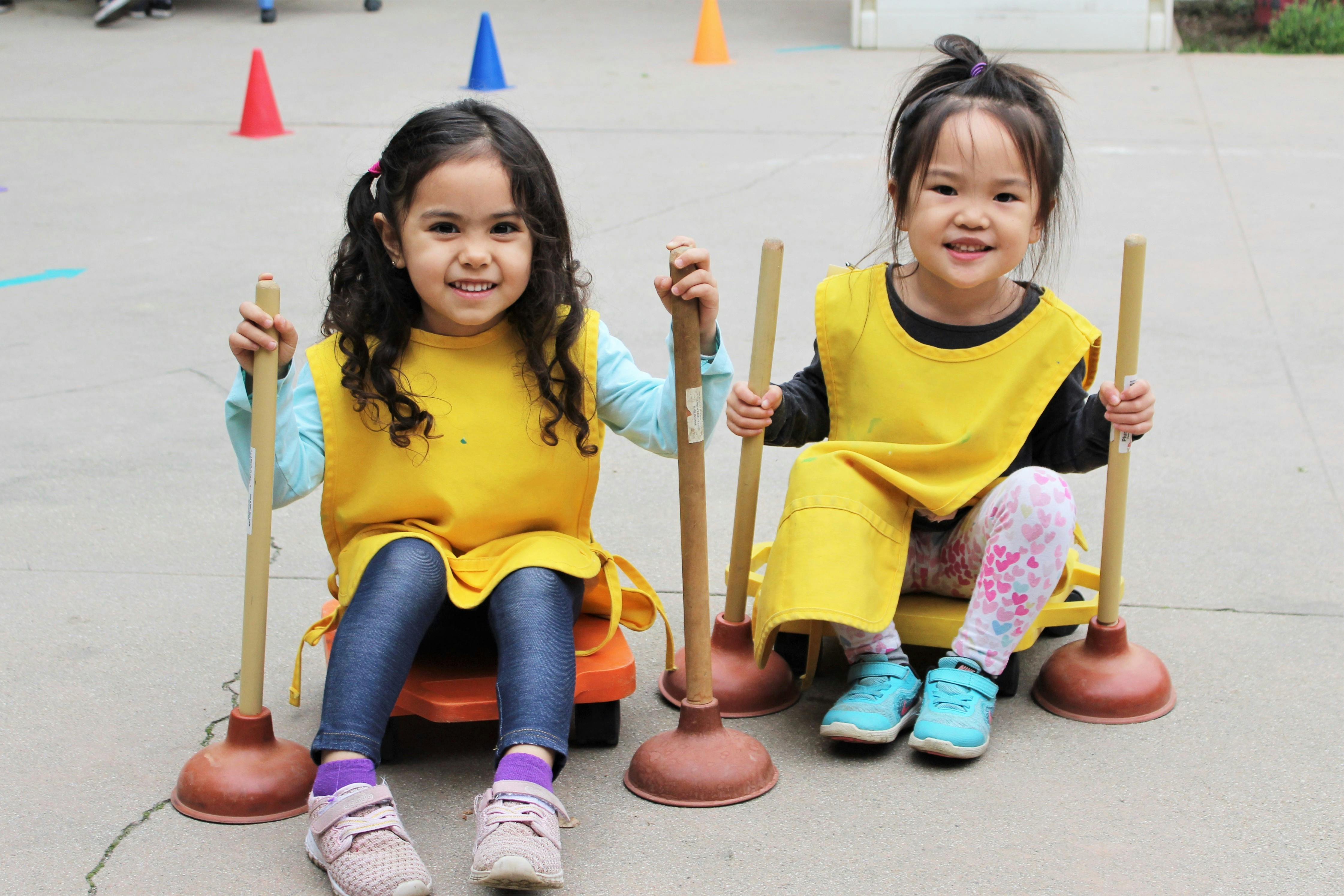 Two children wearing yellow aprons, sitting on small carts and holding plungers
