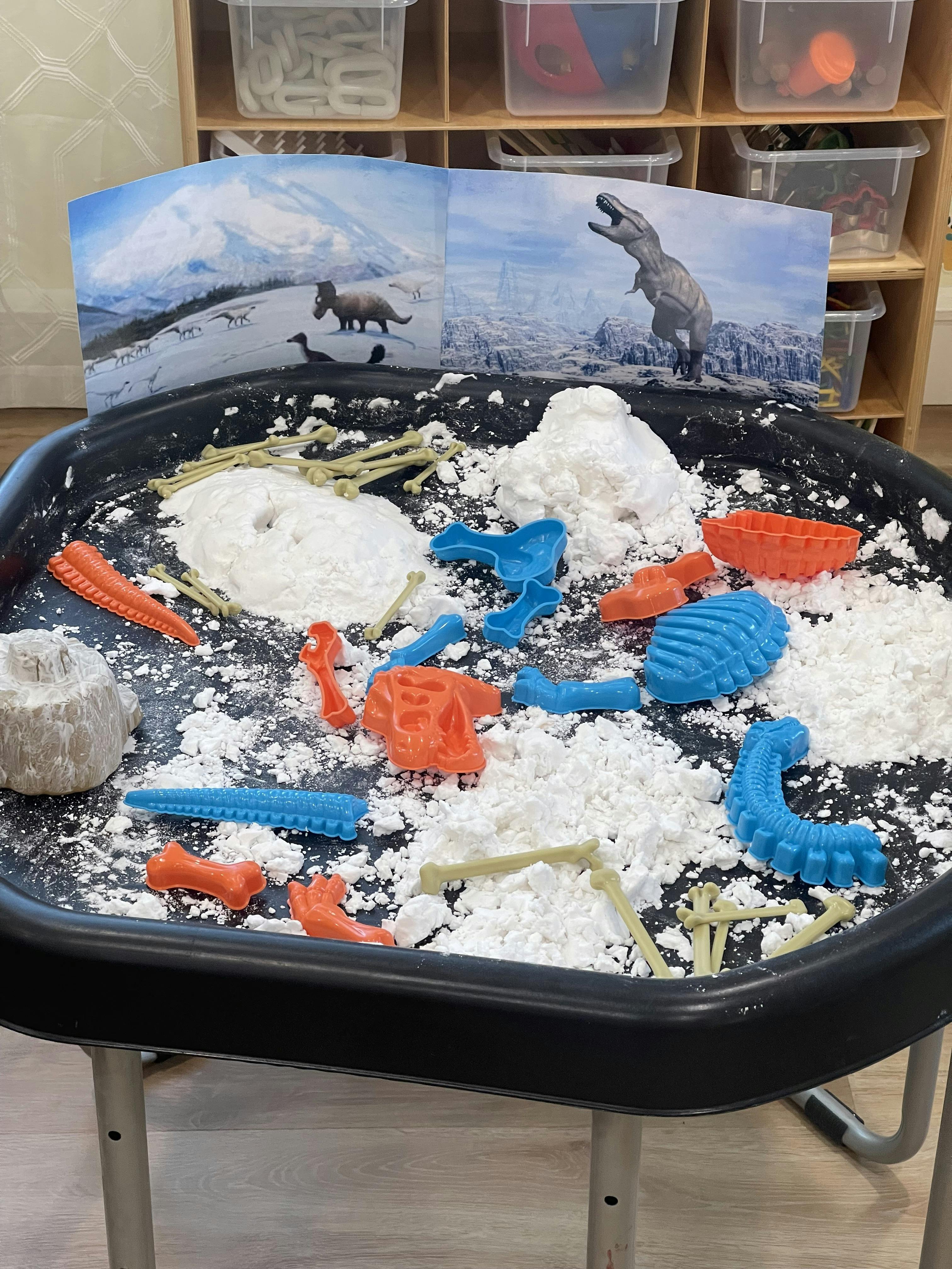 Dinosaur-themed sensory bin with white powder, plastic bones, and tools