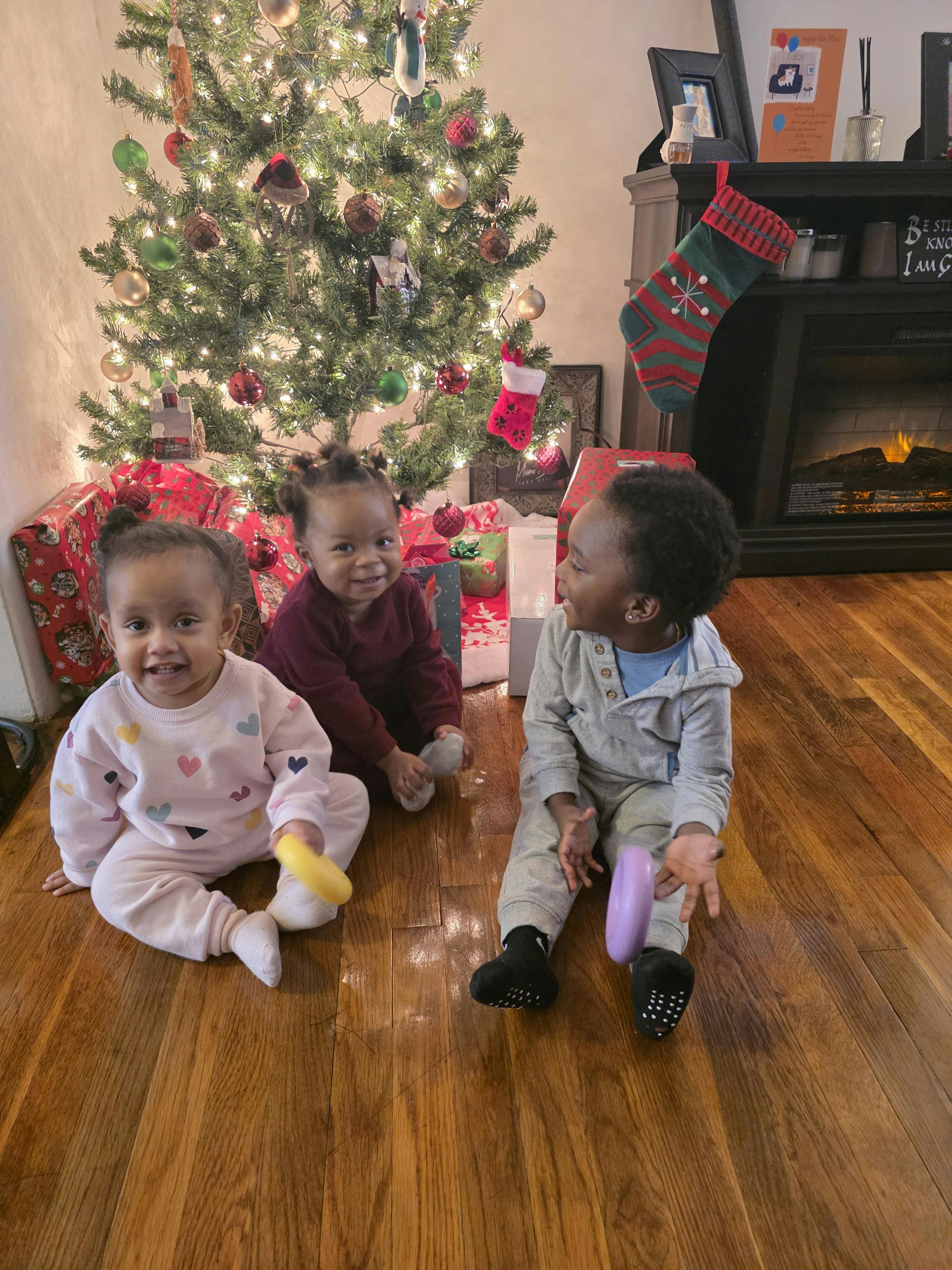 Three children sitting on a wooden floor in front of a Christmas tree