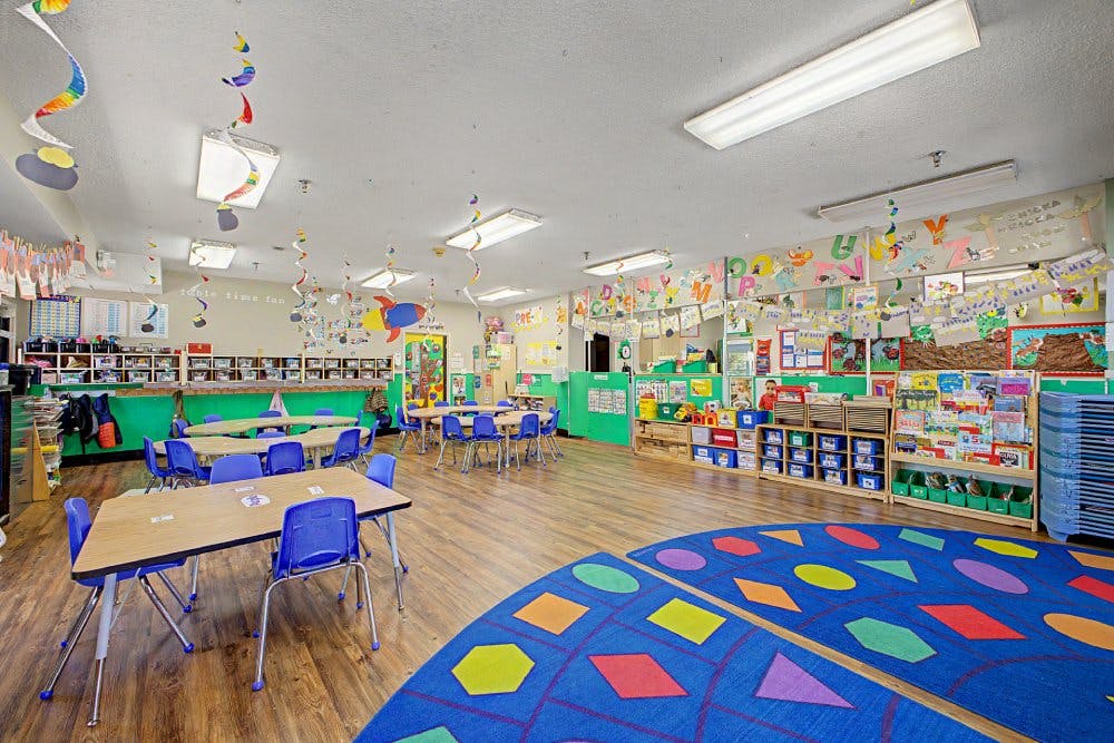 Colorful preschool classroom with child-sized tables and chairs, shelves, and a rug