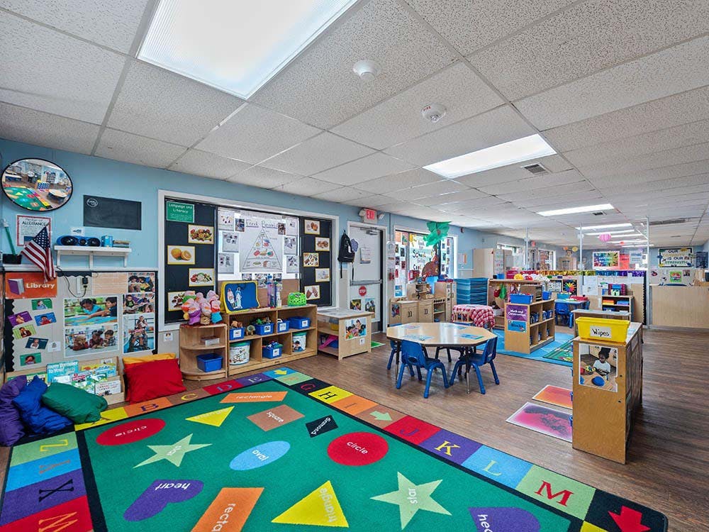 Colorful classroom with a shape rug, tables, chairs, and shelves of learning materials