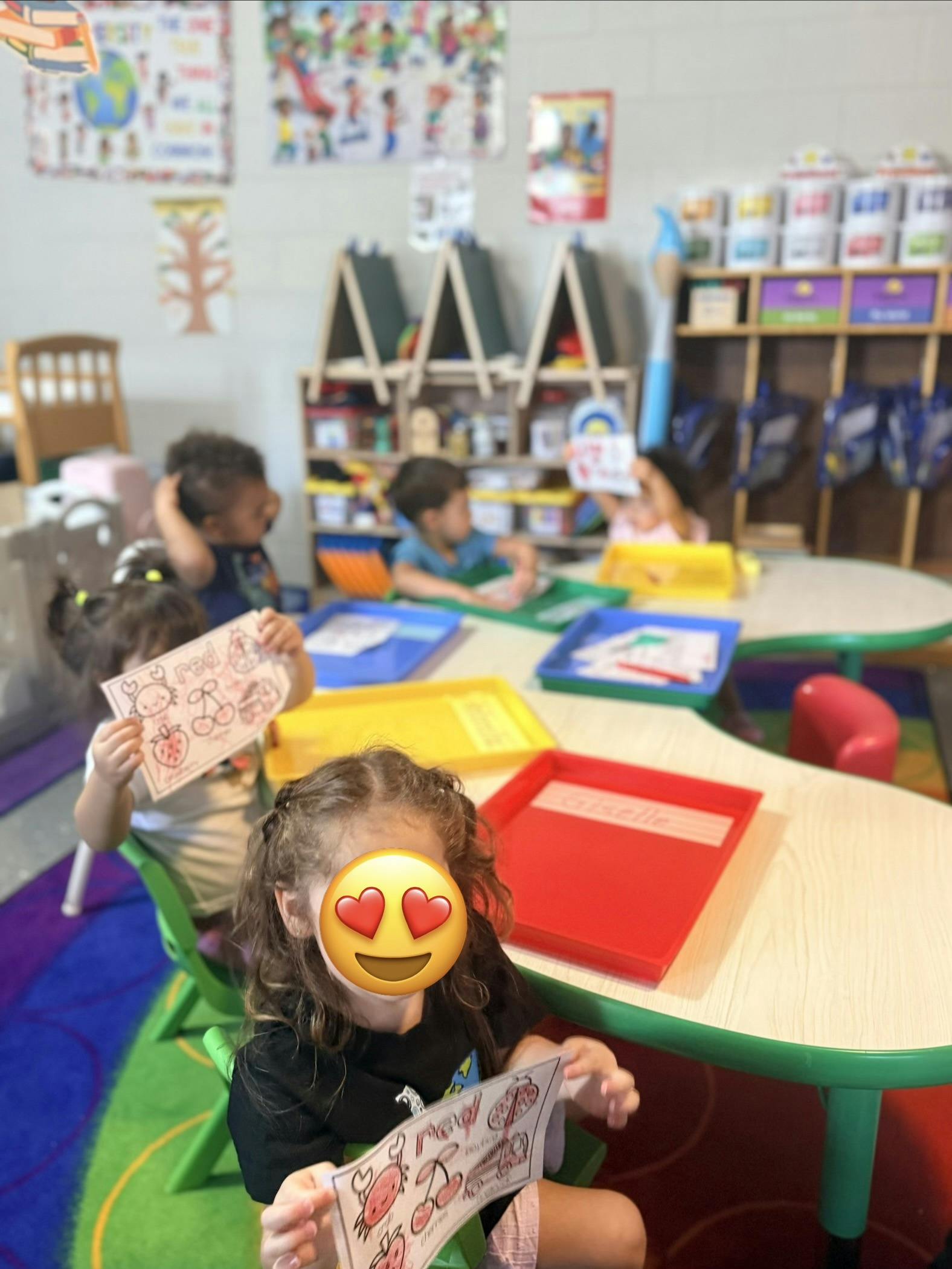 Children sitting at tables in a classroom holding papers with colored drawings
