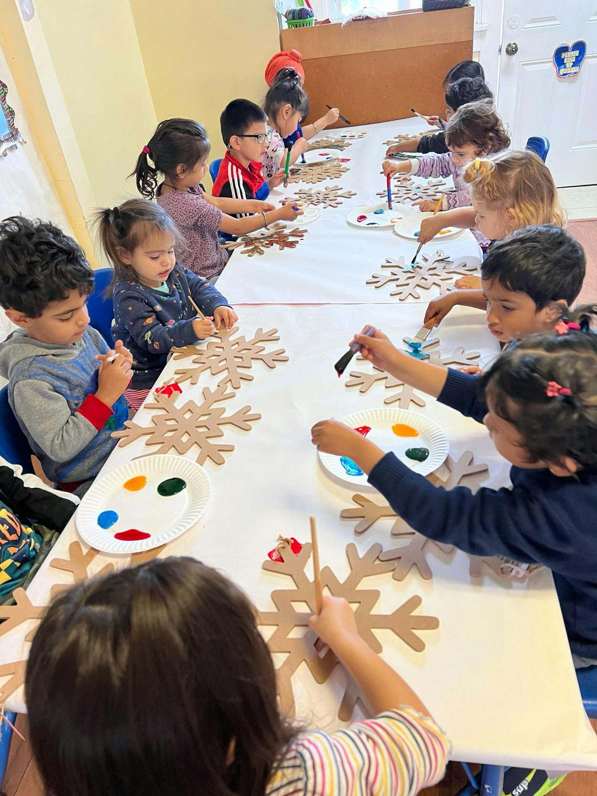 Children painting snowflake cutouts at a long table in a classroom