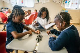 Three children at a table, one reading a book