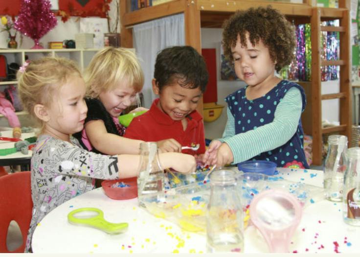 Four children playing with colorful materials at a table in a classroom