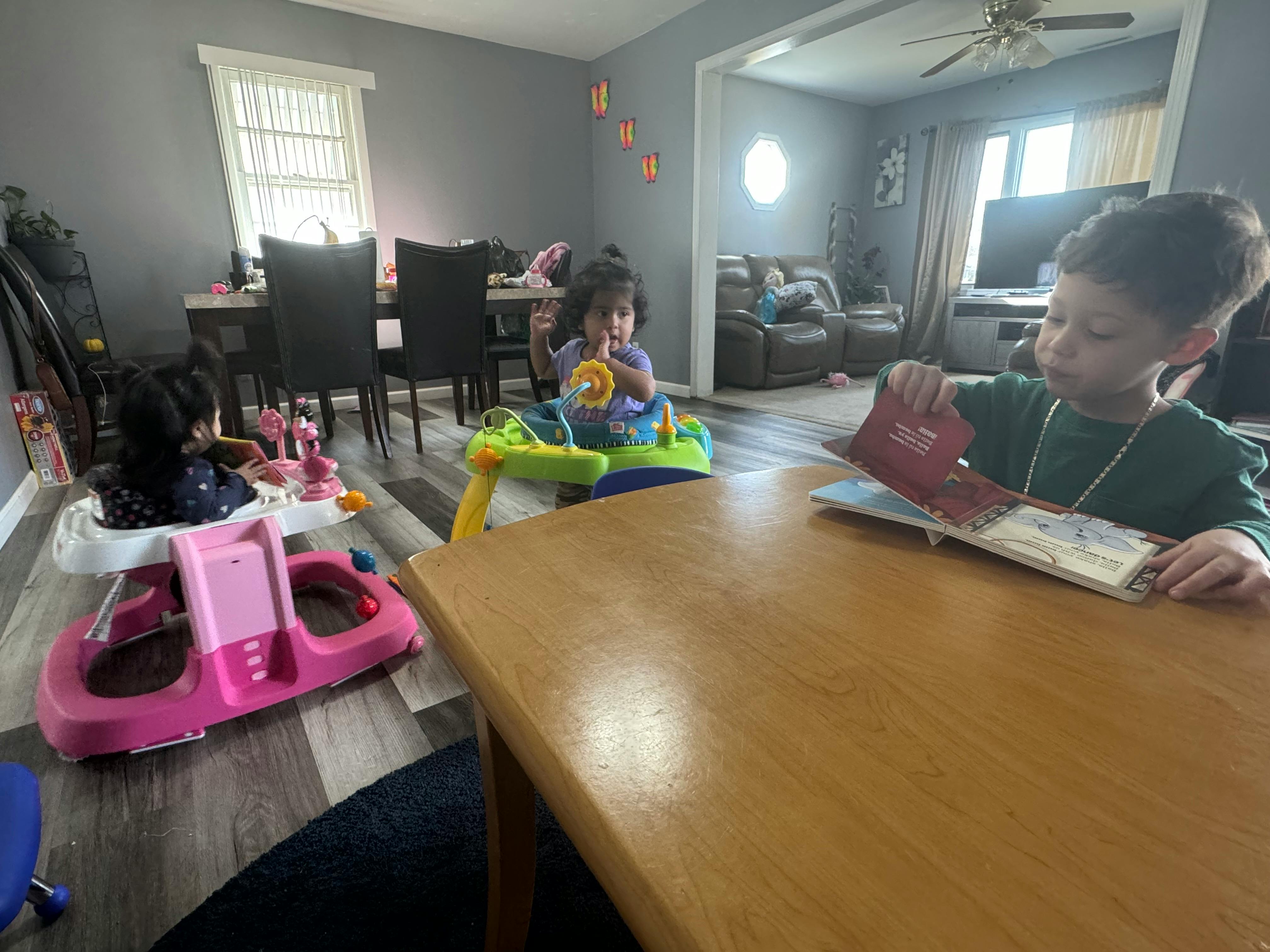 Child reading a book at a table, another child in a baby walker