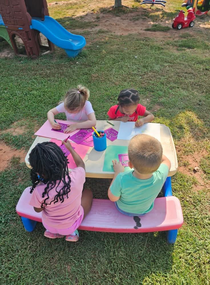Four children coloring at a small picnic table in a grassy outdoor area