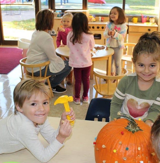 Children and an adult in a classroom, a child holds a toy hammer near a pumpkin