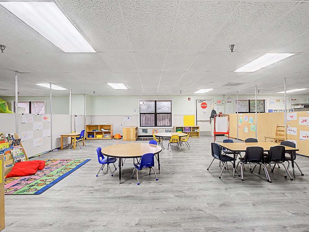 Classroom with small tables and chairs, a colorful rug, and child drawings on walls
