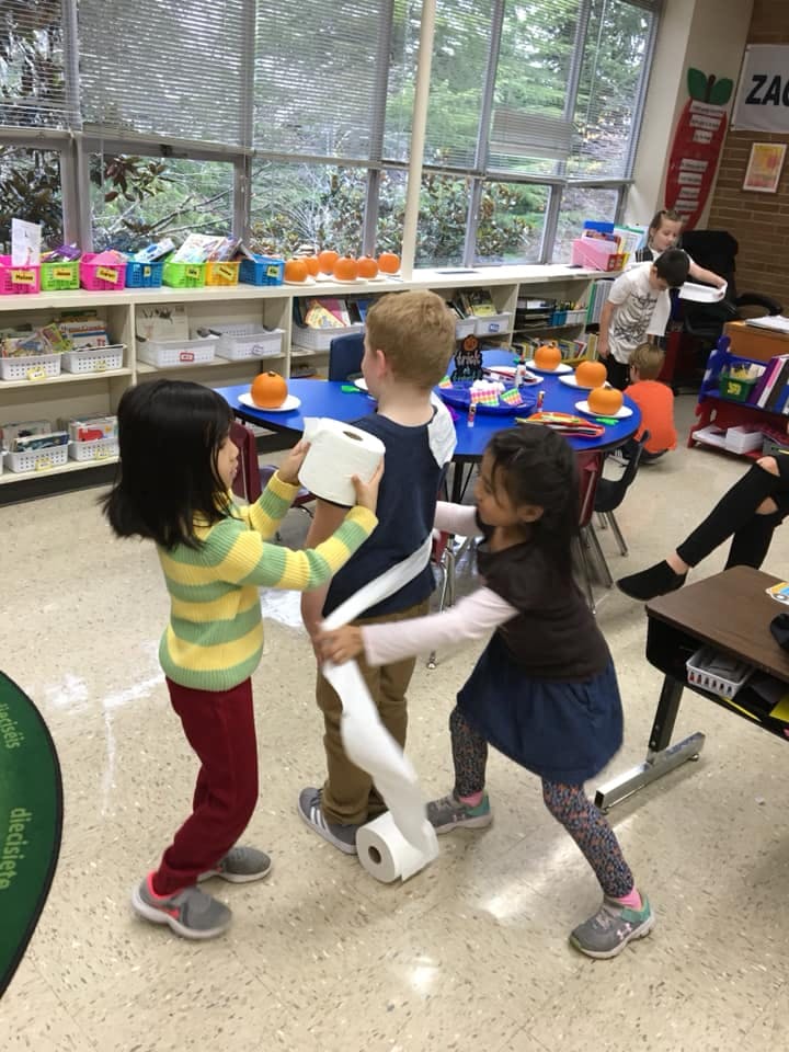 Three children wrapping a fourth child in toilet paper in a classroom