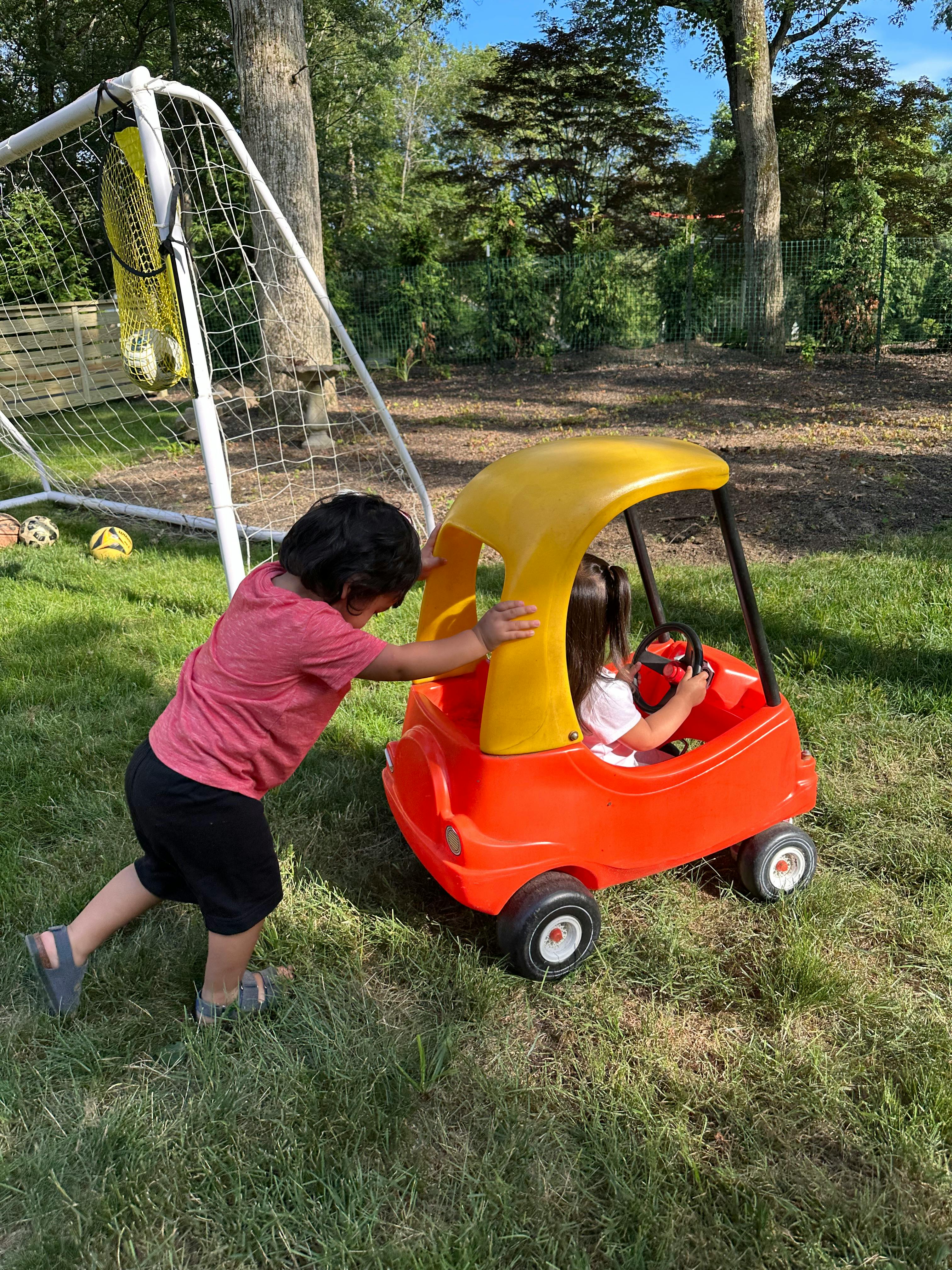 Child in a red and yellow toy car with another child pushing from behind