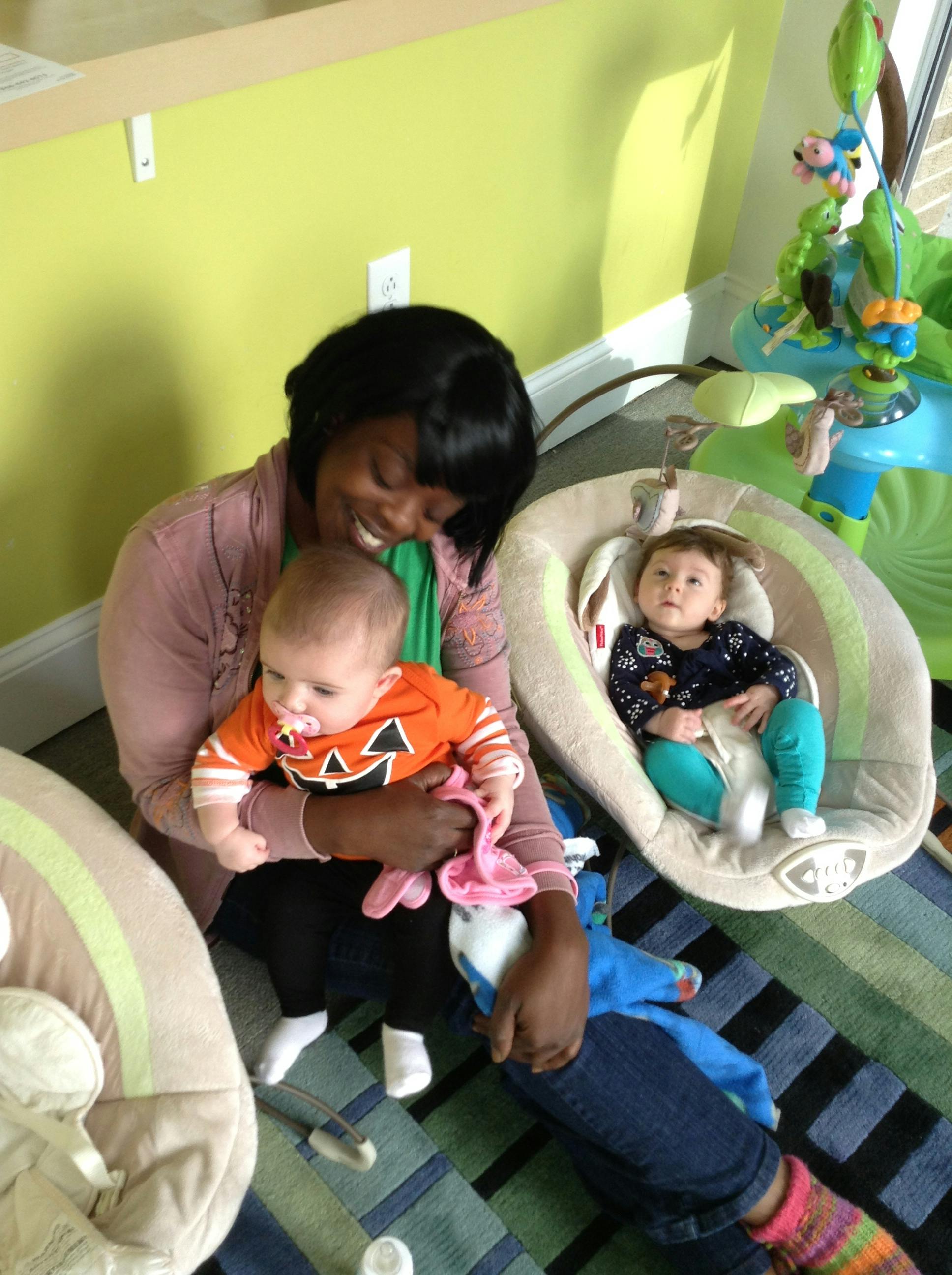 Adult holding a baby while another child rests in a baby seat nearby