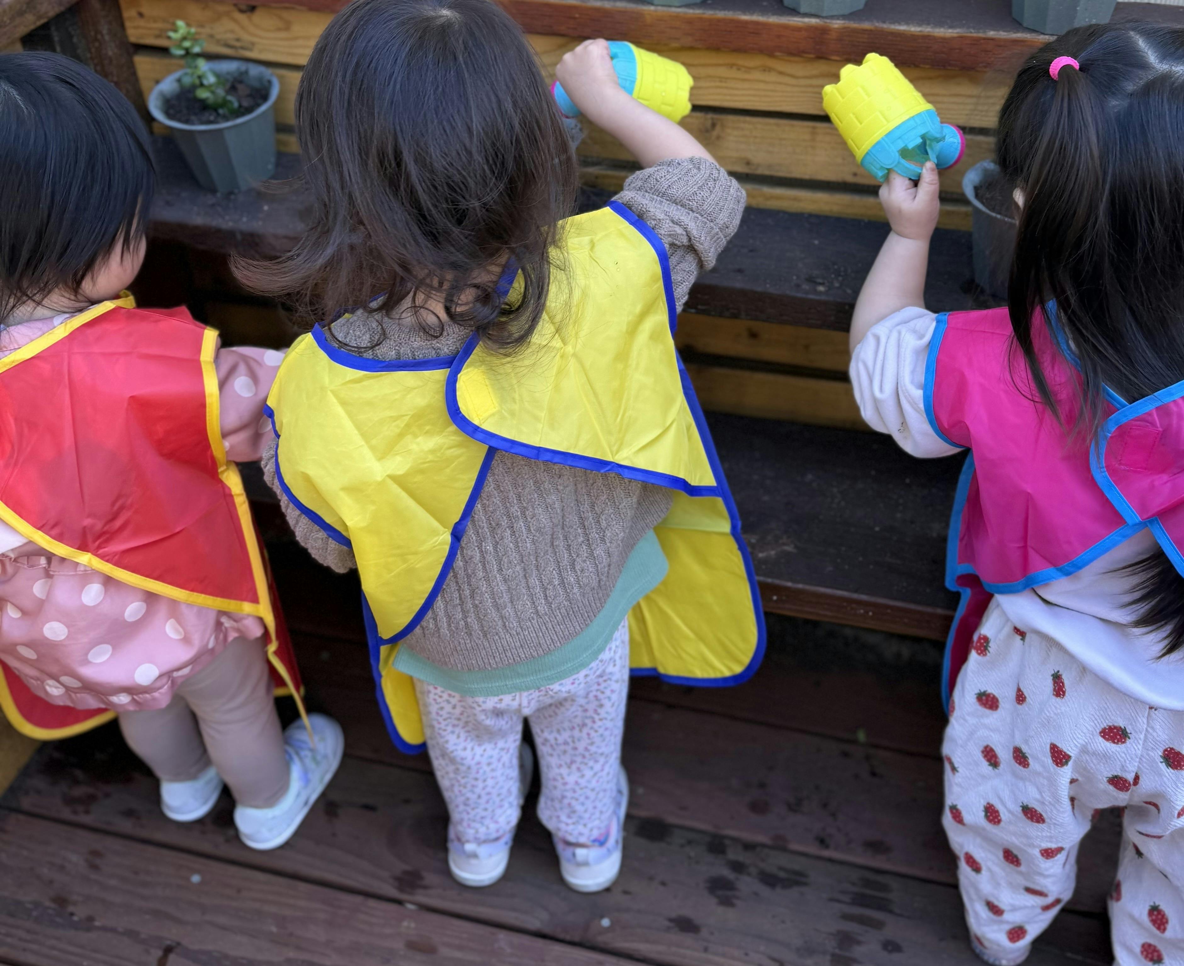 Three children wearing aprons playing with toy watering cans near potted plants