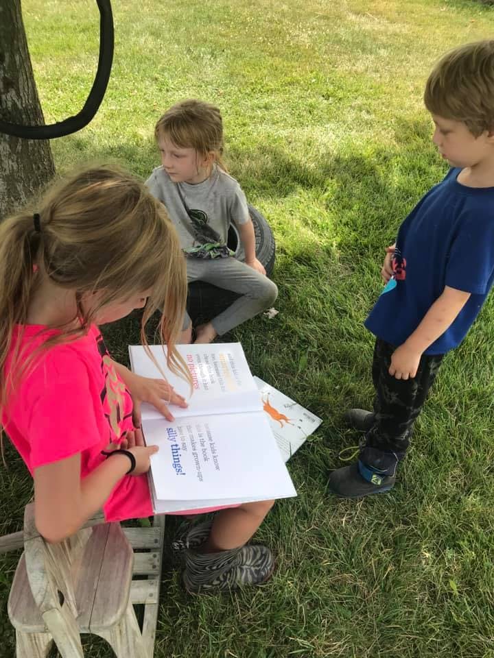 Child in pink shirt reading a book outdoors with two other children nearby