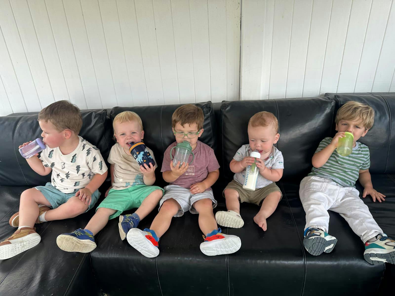 Five children sitting on a black couch drinking from bottles and sippy cups