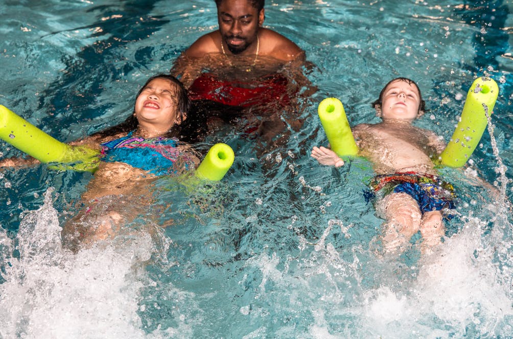 Person in a pool with two children using pool noodles