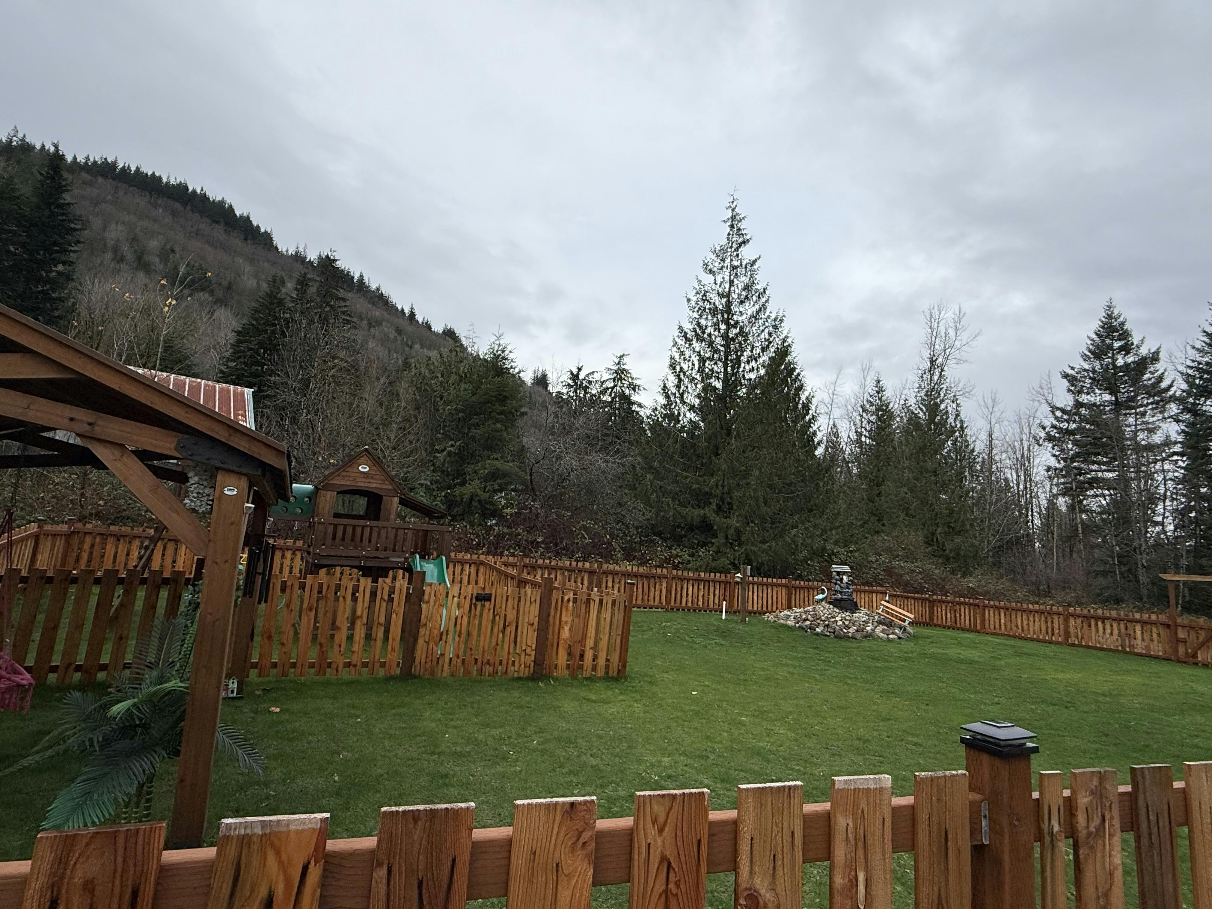 backyard with a wooden play structure and fence, backed by a forest and hill under a cloudy sky