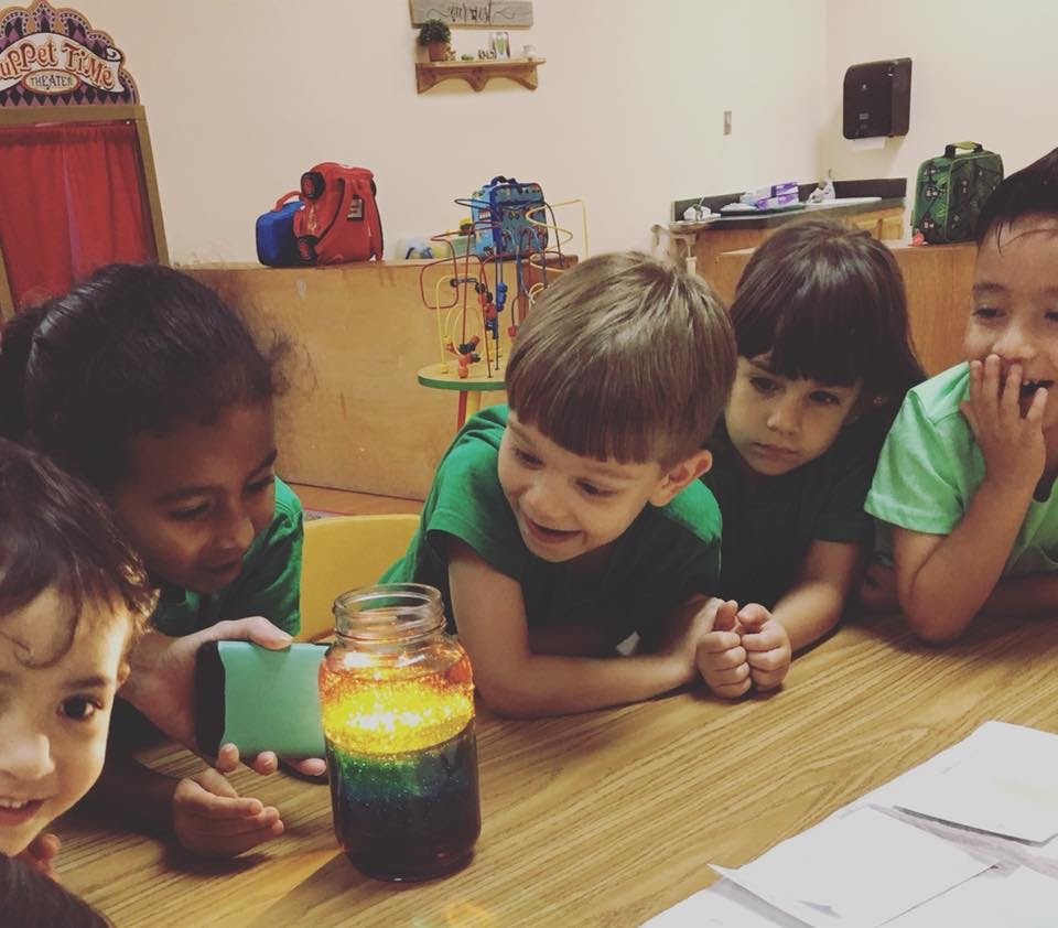 Children looking at a colorful jar on a table in a classroom