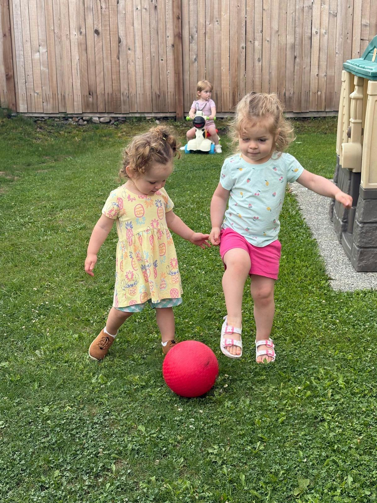 Two children playing with a red ball on grass, another child in background