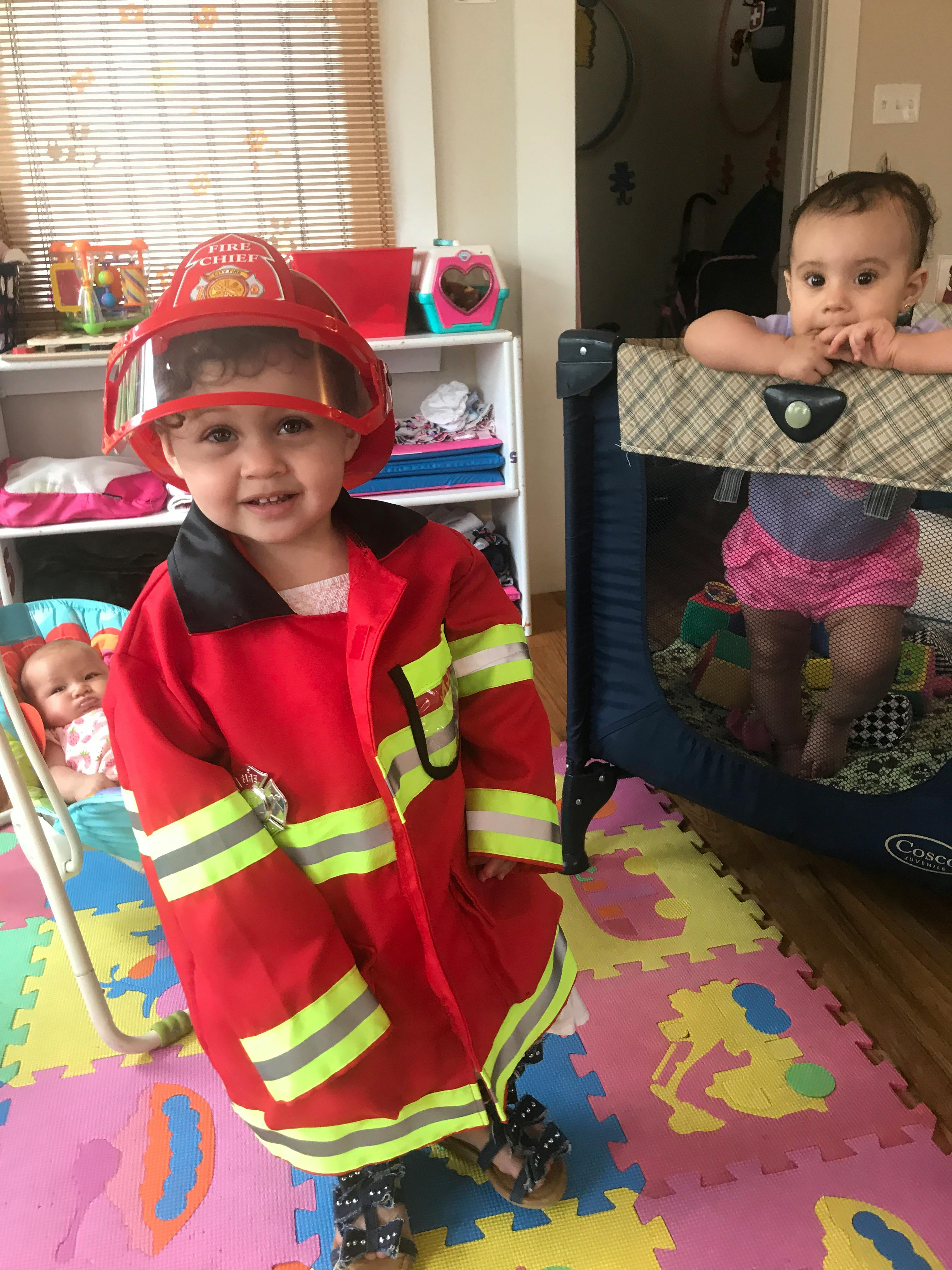 Child in firefighter costume standing, another child in playpen
