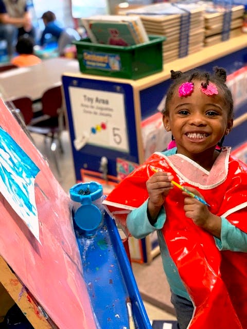 Child in a red smock painting at an easel in a classroom
