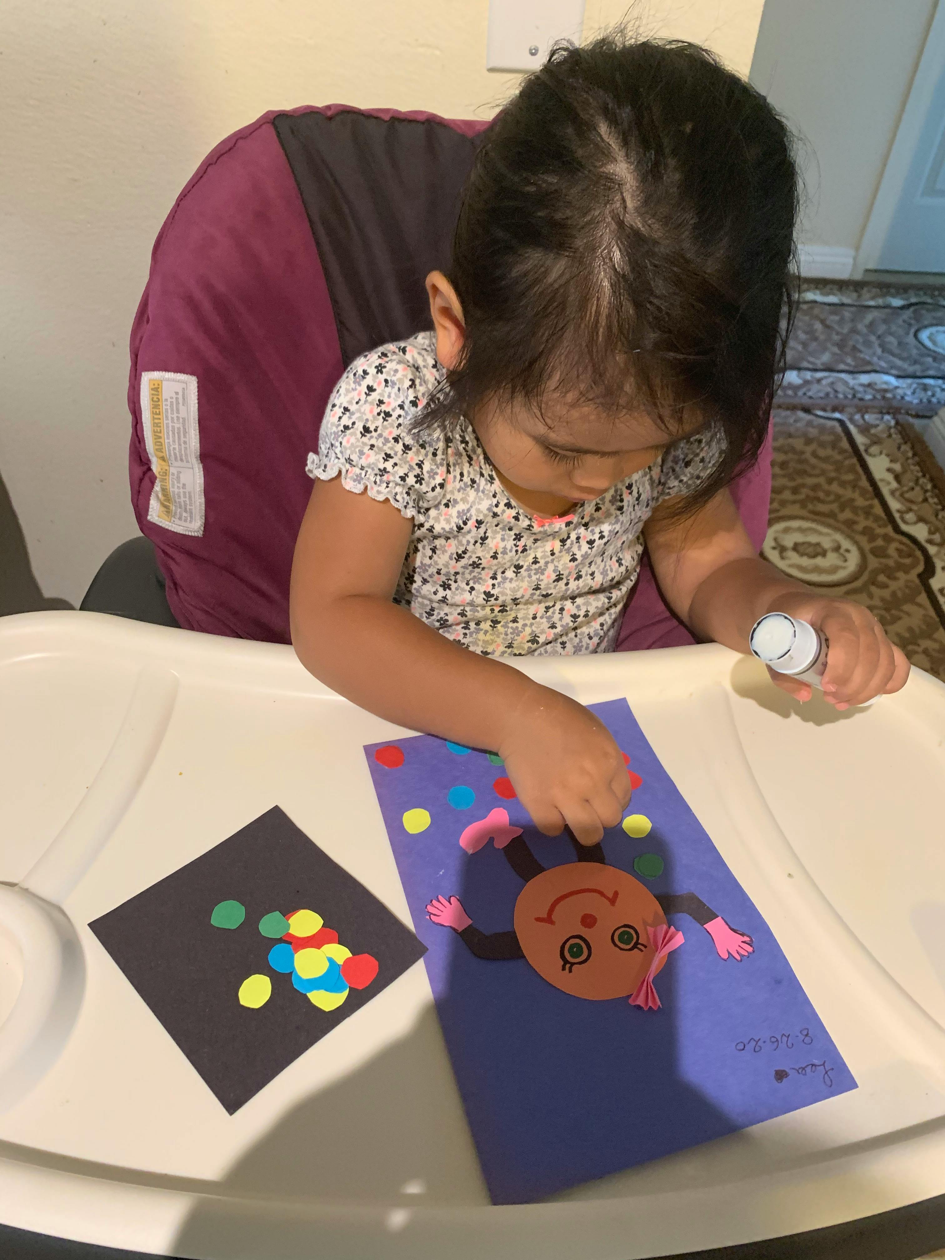 Child gluing paper cutouts onto a purple sheet of paper