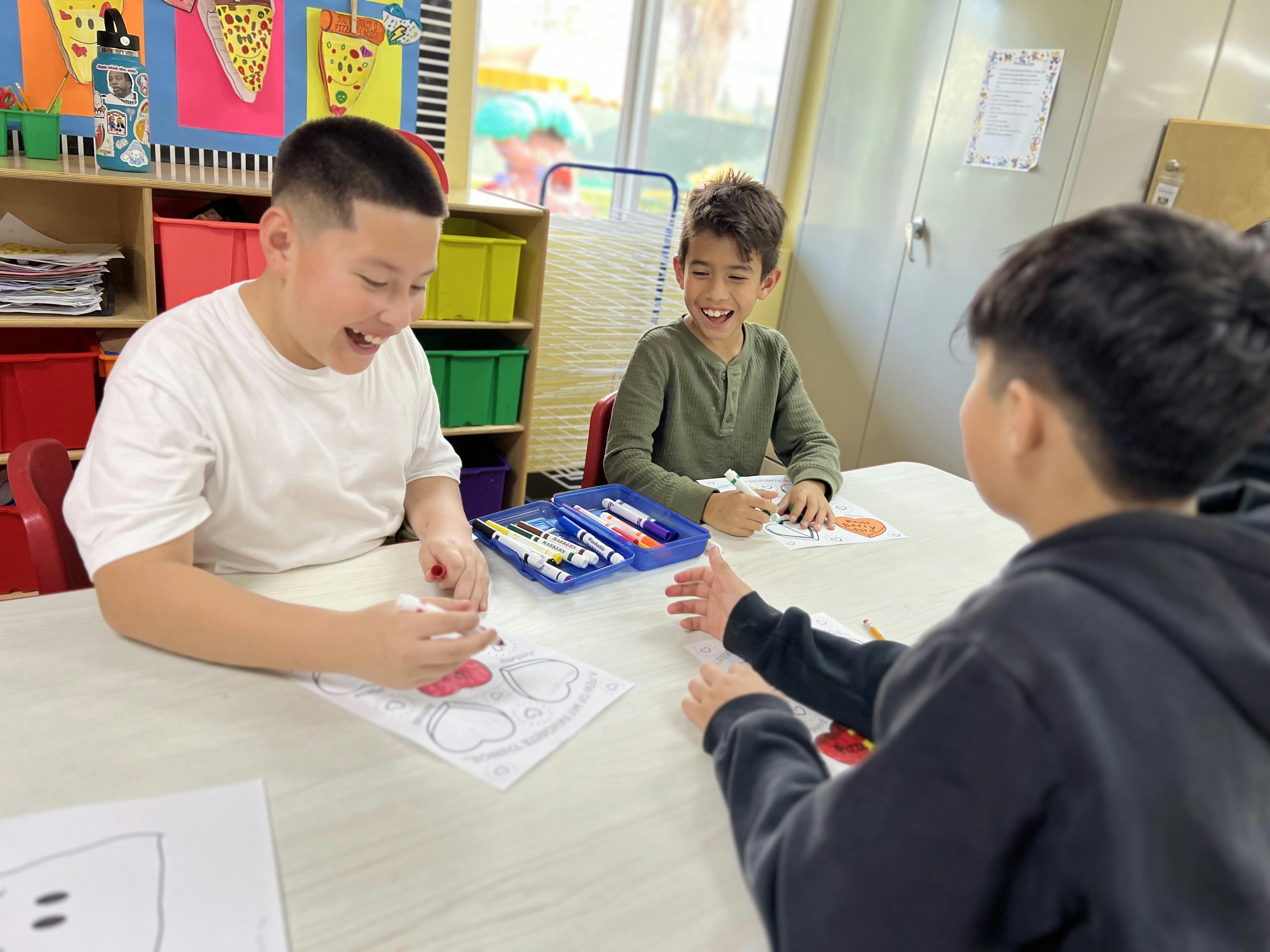 Three children sitting at a table, drawing and interacting in conversation