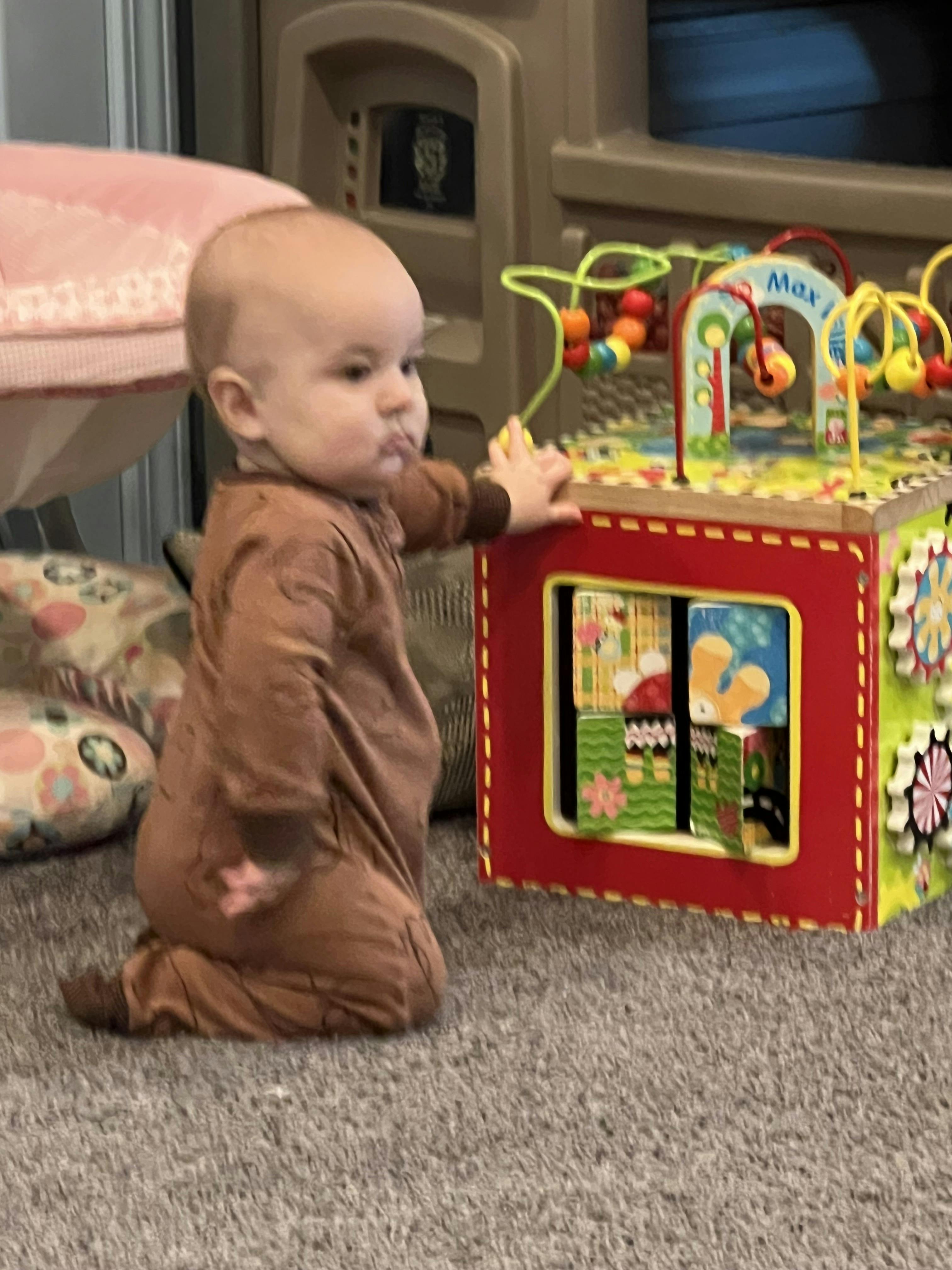 Baby in a brown outfit on knees, holding a toy activity cube