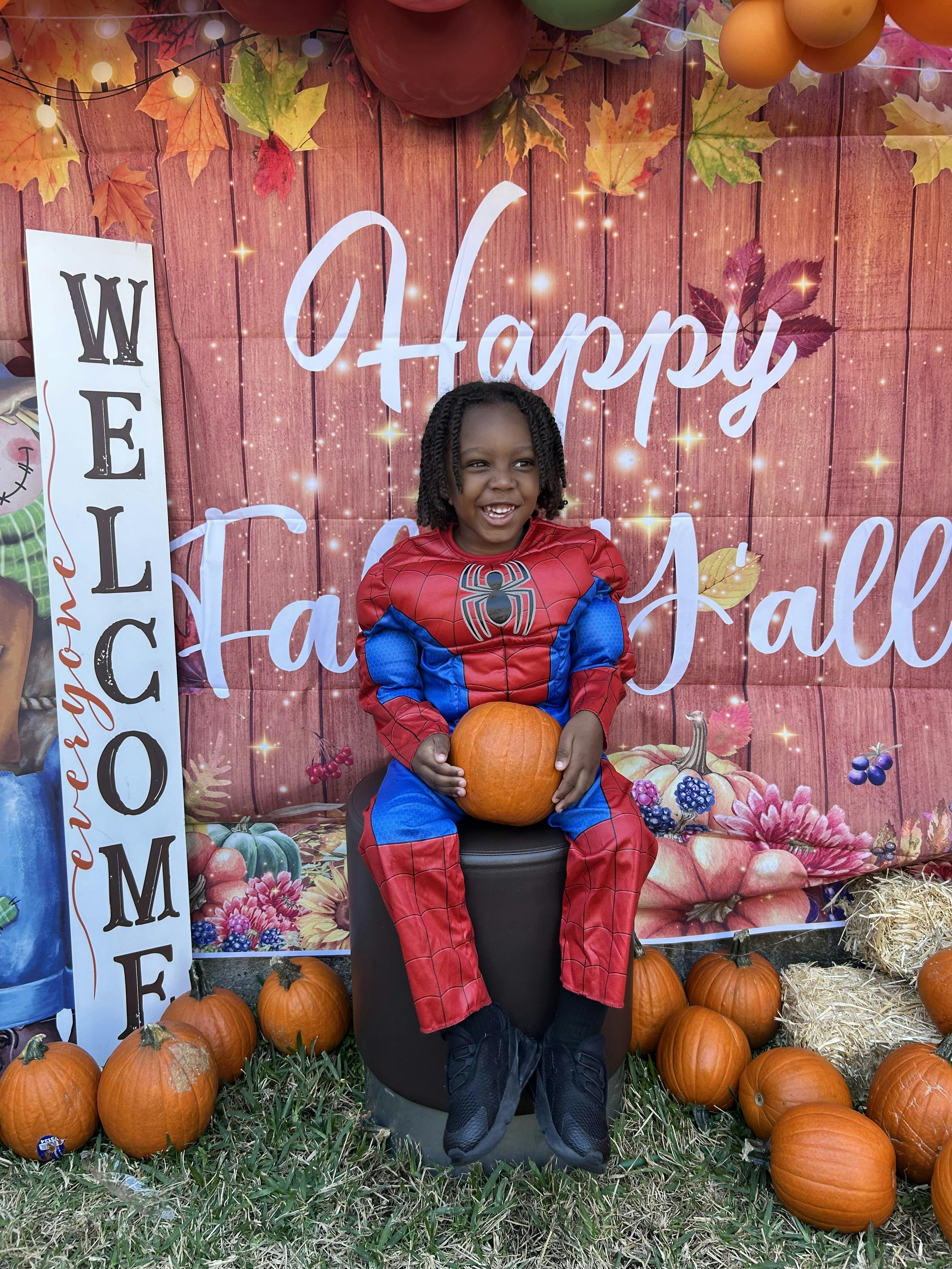 Child in a Spider-Man costume holding a pumpkin, in front of a "Happy Fall" backdrop