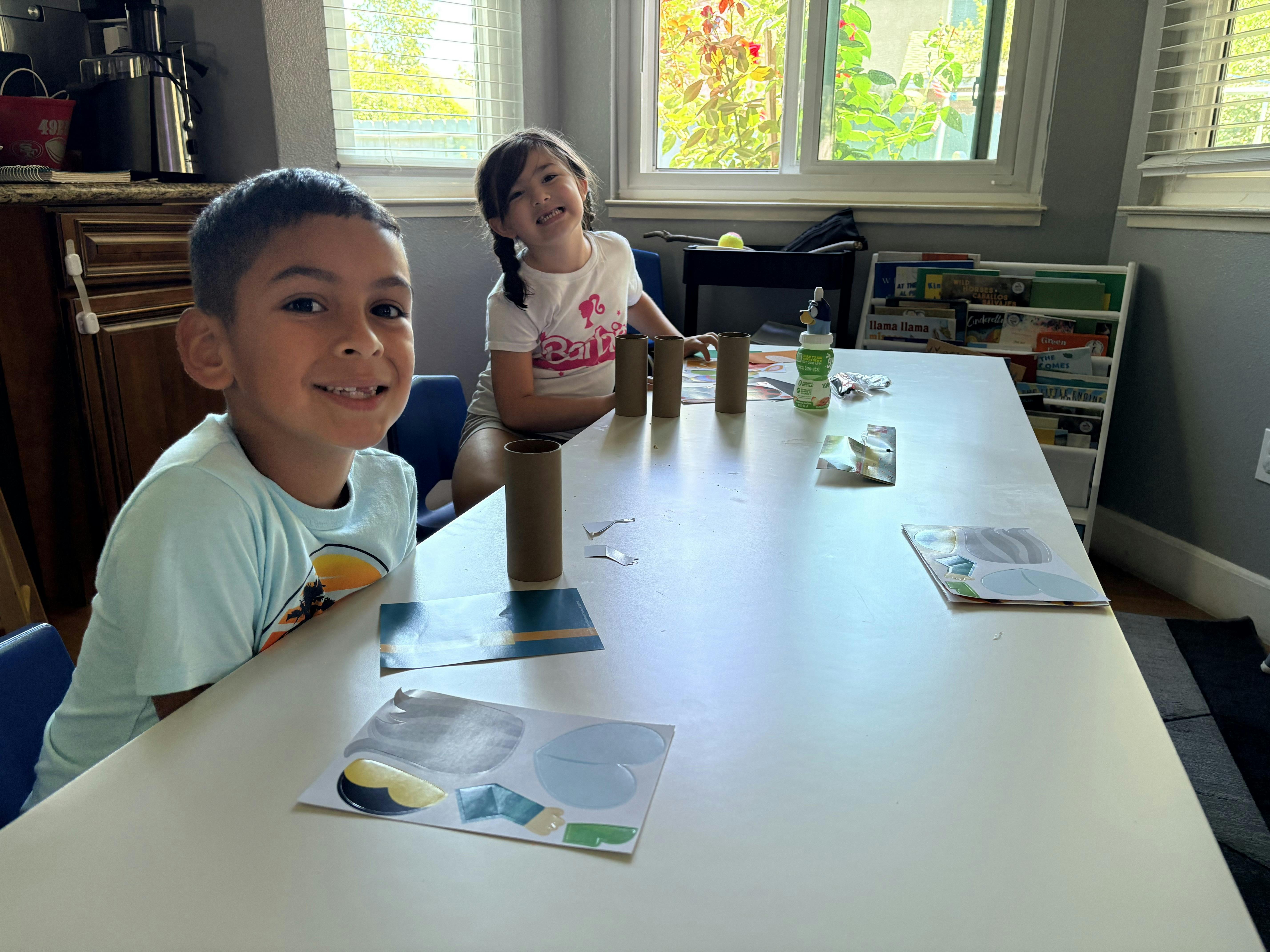 Two children sitting at a table crafting with cardboard tubes and paper cutouts