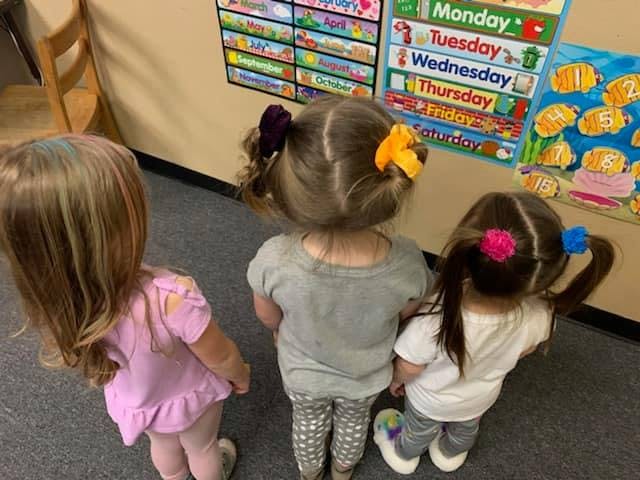 Three children standing with their backs to the camera in a classroom