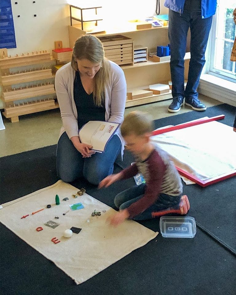 Person kneeling with a book watching a child playing with objects on a mat