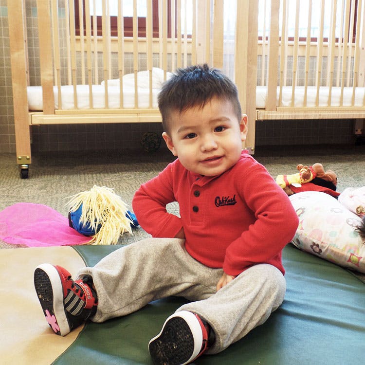 Child sitting on a mat in a room with cribs