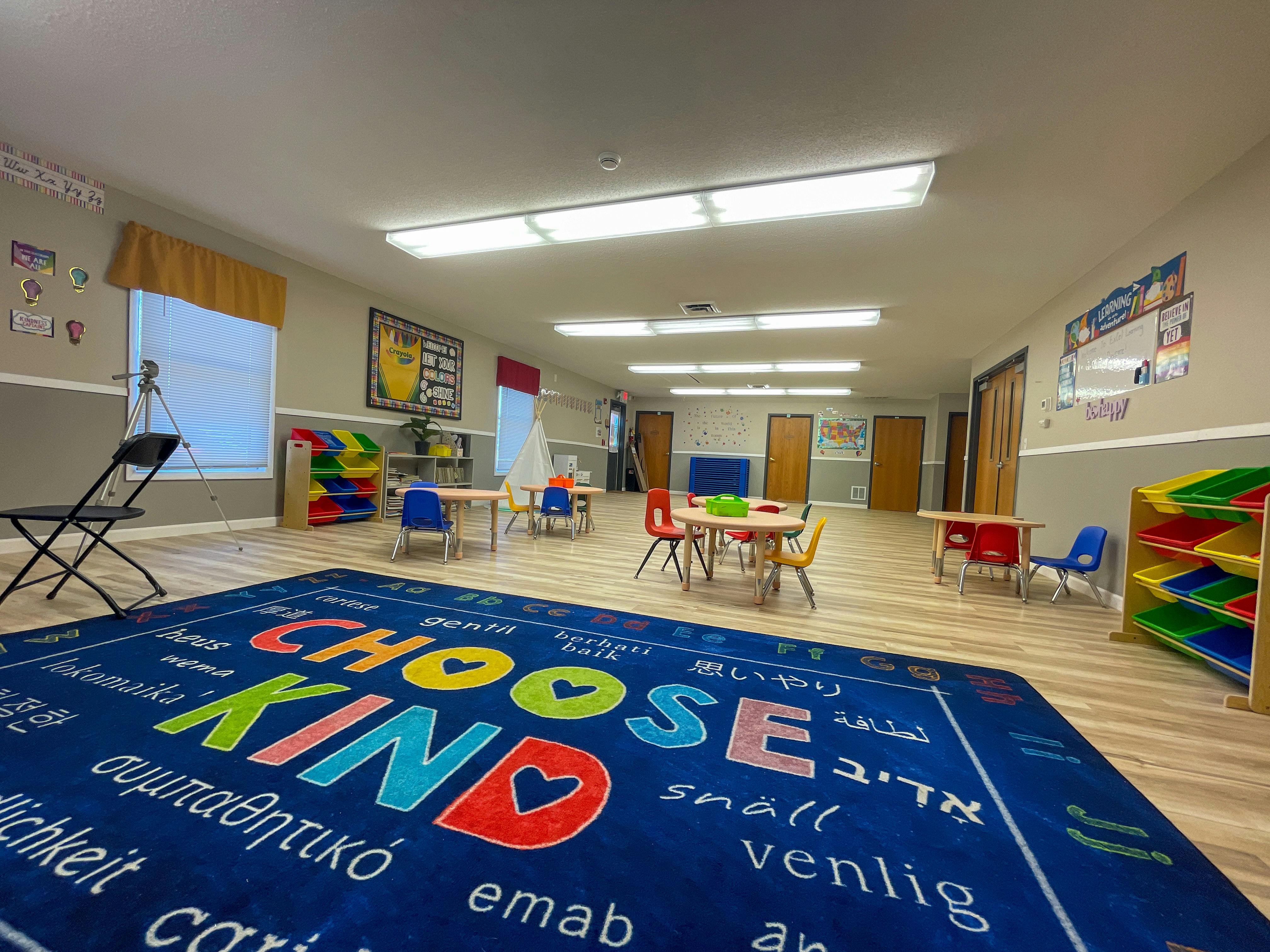 Classroom with small tables and chairs, and a blue rug with "CHOOSE KIND"