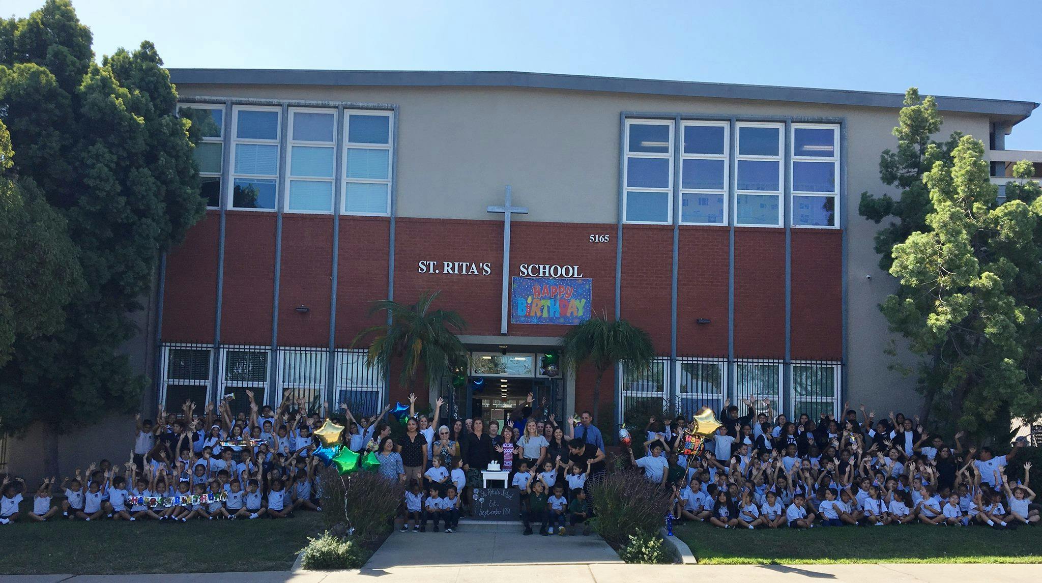 Large group of people gathered in front of a school building with "St. Rita's School" sign