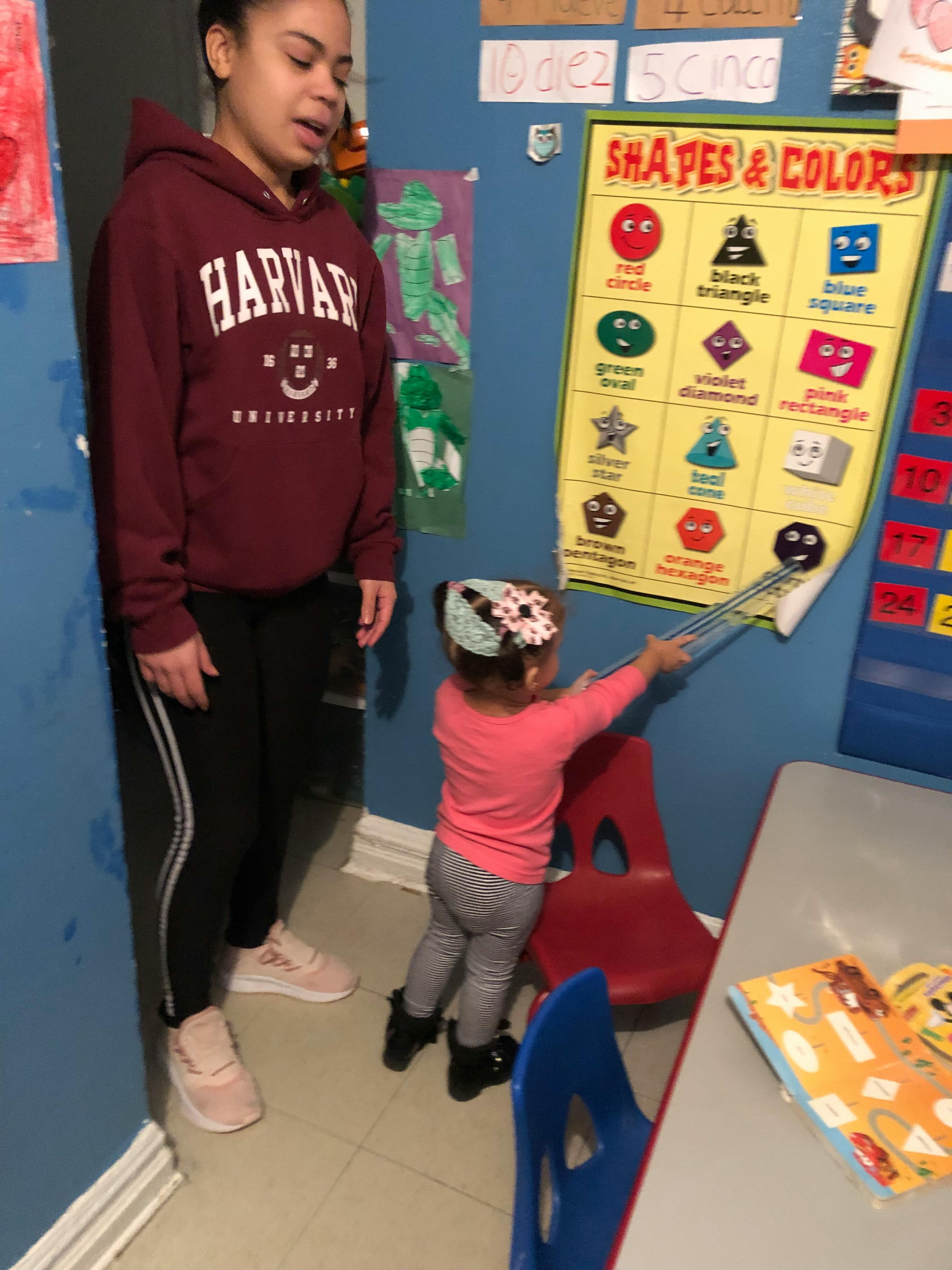 Person and child in a classroom, child pointing at a shapes and colors chart